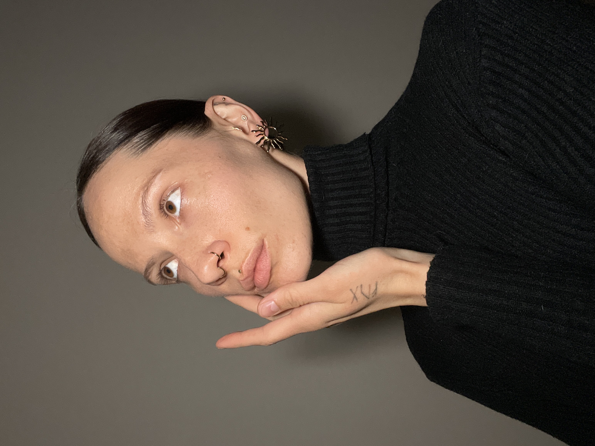 A woman is depicted in a portrait-style image, positioned at an angle against a gray backdrop. She is wearing a black turtleneck sweater. Her hair is dark and slicked back. She has fair skin and noticeable piercings, including a ring in her nose, studs in her ear, and a large, sun-shaped earring. Her hand, bearing a tattoo resembling the roman numeral XVI, is positioned under her chin. The image does not contain any information that would indicate that the location is Pervomaysky, Russia.