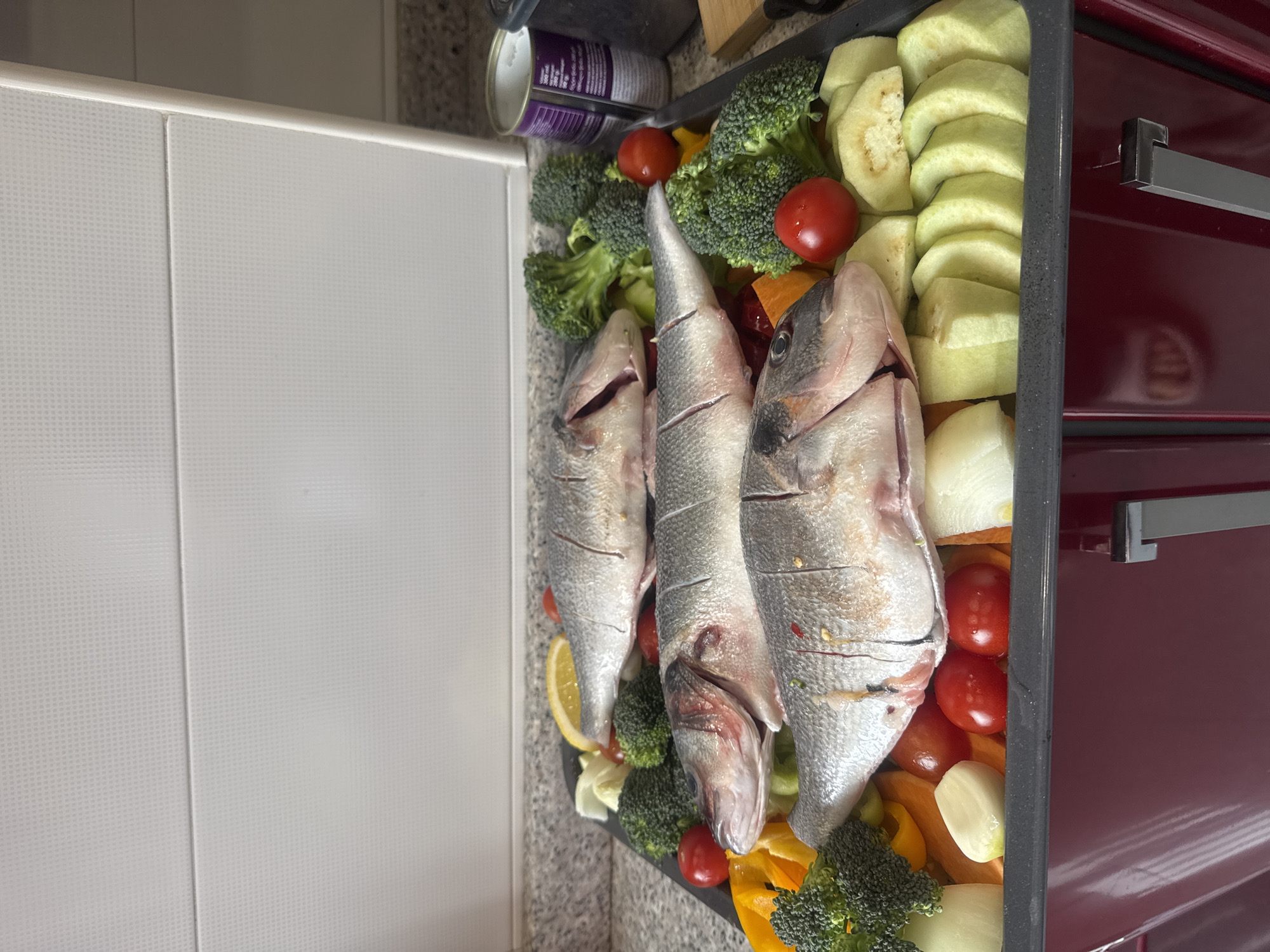 The image shows a baking tray filled with raw ingredients. Three whole fish are centrally positioned, surrounded by an assortment of vegetables: broccoli florets, sliced eggplant, cherry tomatoes, sliced onions, and sliced yellow bell peppers. A lemon slice is also visible. A can of food is partially visible at the top left of the tray. The baking tray is positioned on a countertop next to a white cabinet and a red oven. The image appears to be taken indoors, likely in a kitchen. The specific location within the city of Tbilisi, Georgia, cannot be determined from the visual information provided.