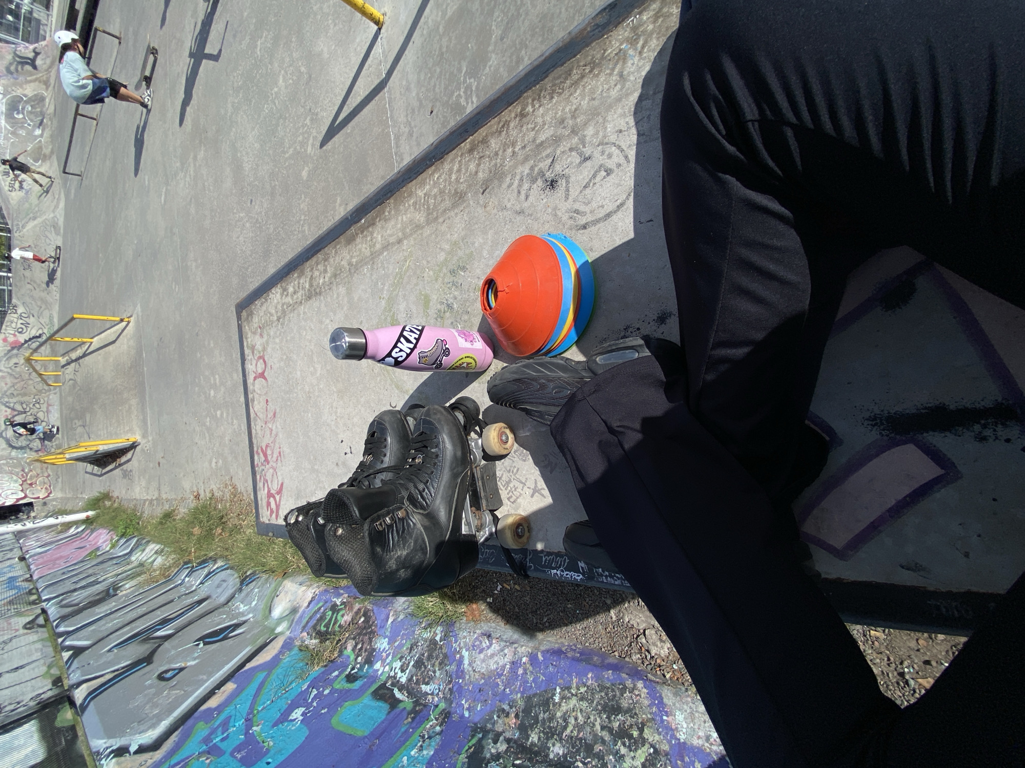 The image is taken at a skatepark in Monte Grande, Argentina. In the foreground, a person wearing black pants and black shoes is sitting on the edge of the graffiti-covered skatepark bowl. Next to the person is a pair of black roller skates resting on a skateboard, a pink water bottle with stickers, and a colorful orange and blue cone. In the background, there are other people skating in the park, with yellow rails and various graffiti designs on the concrete surfaces. The overall environment suggests an outdoor, recreational space.