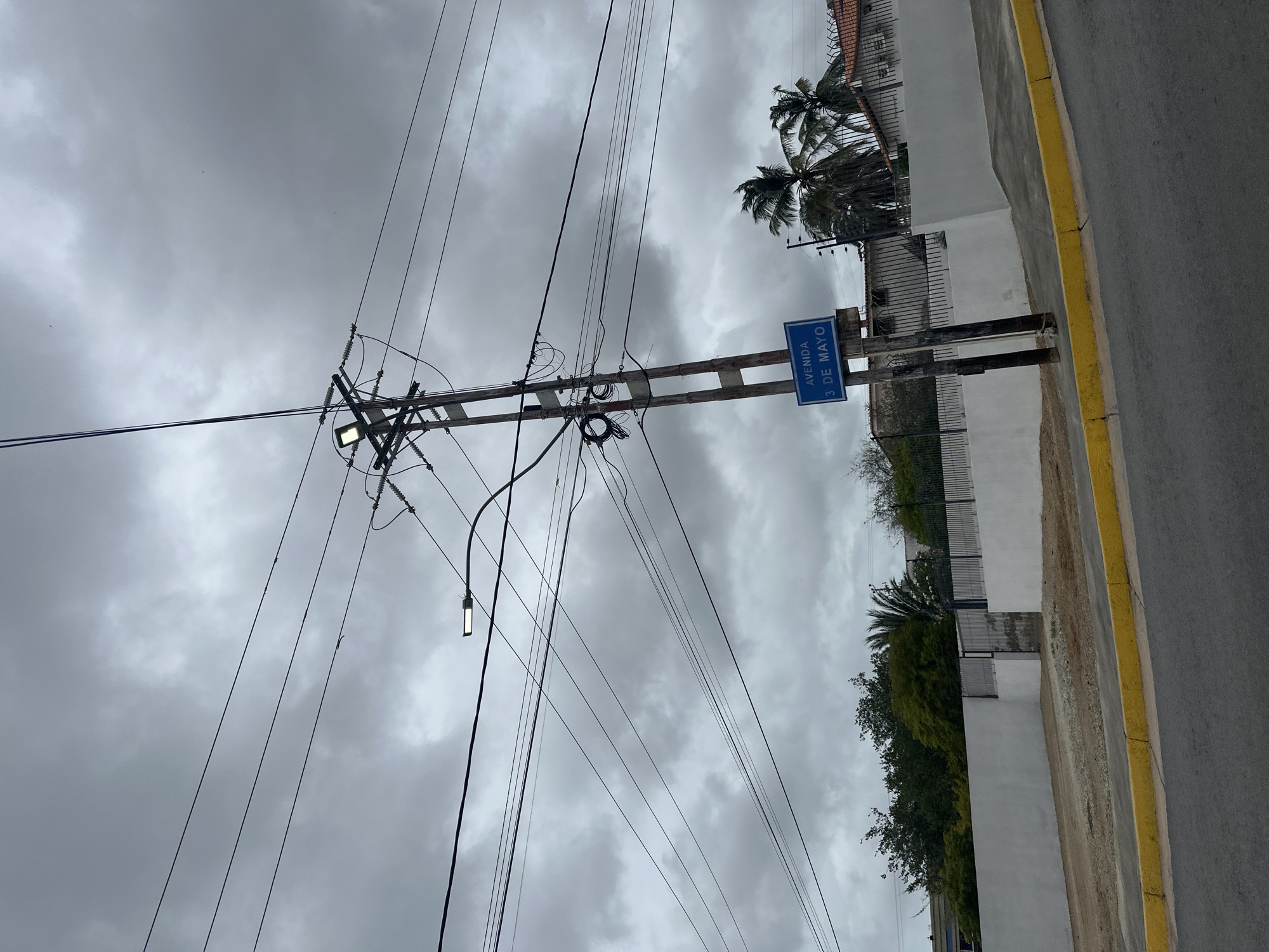 The image depicts a street scene in Juan Griego, Venezuela, under a cloudy sky. A wooden utility pole with several electrical wires and a street lamp stands prominently in the foreground. A blue street sign reading "AVENIDA 3 DE MAYO" is attached to the pole. In the background, a white wall stretches across the frame, with a black metal gate in the center. Lush green trees and palm trees are visible behind the wall, along with a red-tiled roof of a building. The street in the foreground is paved, with a yellow curb bordering it. No people are visible in the image.