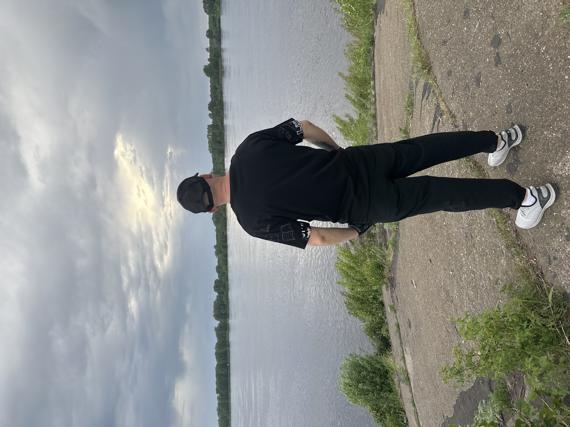 A photograph, taken from a high vantage point, shows a person standing next to a body of water. The person is facing away from the camera, wearing a black shirt, black pants, a black baseball cap, and white sneakers. The person is on a concrete surface with green vegetation growing along the edge. Beyond the concrete is a body of water that stretches into the distance, flanked by trees on both sides. The sky is overcast with dark clouds and a small patch of sunlight breaking through. The location is Chaikovsky, Russia.