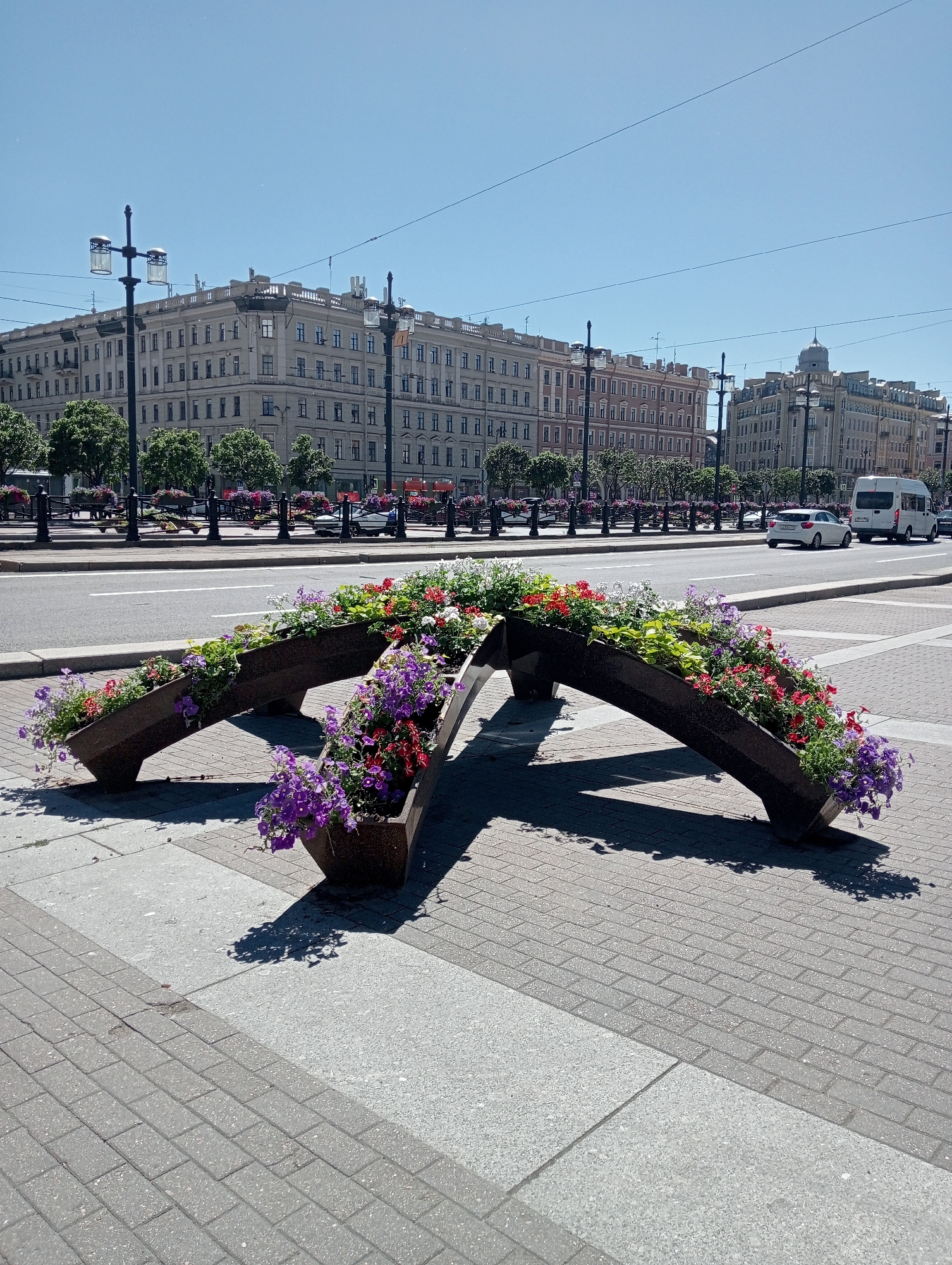 In a bright, sunny daytime scene in Saint Petersburg, Russia, a modern, angular planter filled with vibrant flowers stands on a paved plaza. The planter is made of dark material and has an unusual, segmented design, creating a sense of motion as it arches upwards and then dips. It is filled with a profusion of flowers in shades of purple, pink, red, and white, with lush green foliage interspersed among them.
In the background, a wide road separates the plaza from a row of elegant, multi-story buildings lining a wide boulevard. These buildings feature classical architecture with numerous windows and decorative cornices, characteristic of Saint Petersburg. Trees with full green canopies line the sidewalk in front of the buildings, and ornate lampposts with multiple lights stand along the street. Further down the road, traffic is visible, including a white van and a white car. The sky is a clear, vibrant blue, suggesting fair weather and a warm day. The overall impression is one of a well-maintained urban environment, with the colorful flowers providing a focal point of natural beauty against the backdrop of historic architecture and city life. There is no visible text in the image.