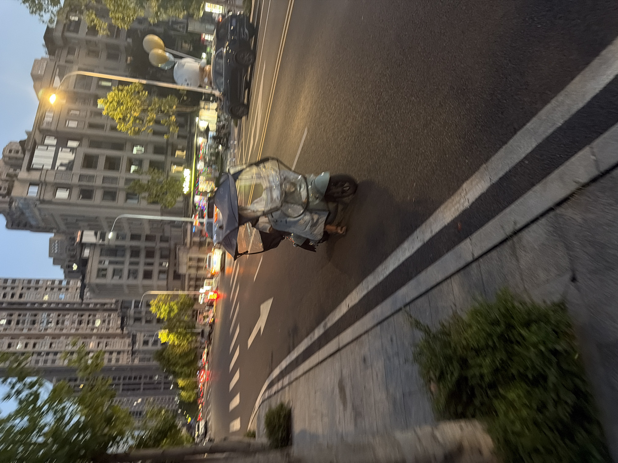 The image depicts a street scene in Jiangjin, China, during twilight. A person is riding a light blue motorized scooter with a clear plastic canopy, a common mode of transport in the area. The scooter is traveling on the right side of a multi-lane road, indicated by white directional arrows painted on the asphalt. Adjacent to the road is a sidewalk with a sparse green bush and a tree trunk on the left. In the background, tall, modern buildings dominate the cityscape, with some windows illuminated. Streetlights are visible, casting a warm glow on the scene. Other vehicles, including cars and what appear to be traffic lights, are present on the road, suggesting urban activity. A large, whimsical balloon sculpture, possibly a character, is visible on the right side of the street. The overall lighting suggests it is either early morning or late evening, with ambient light from the sky and artificial sources.
