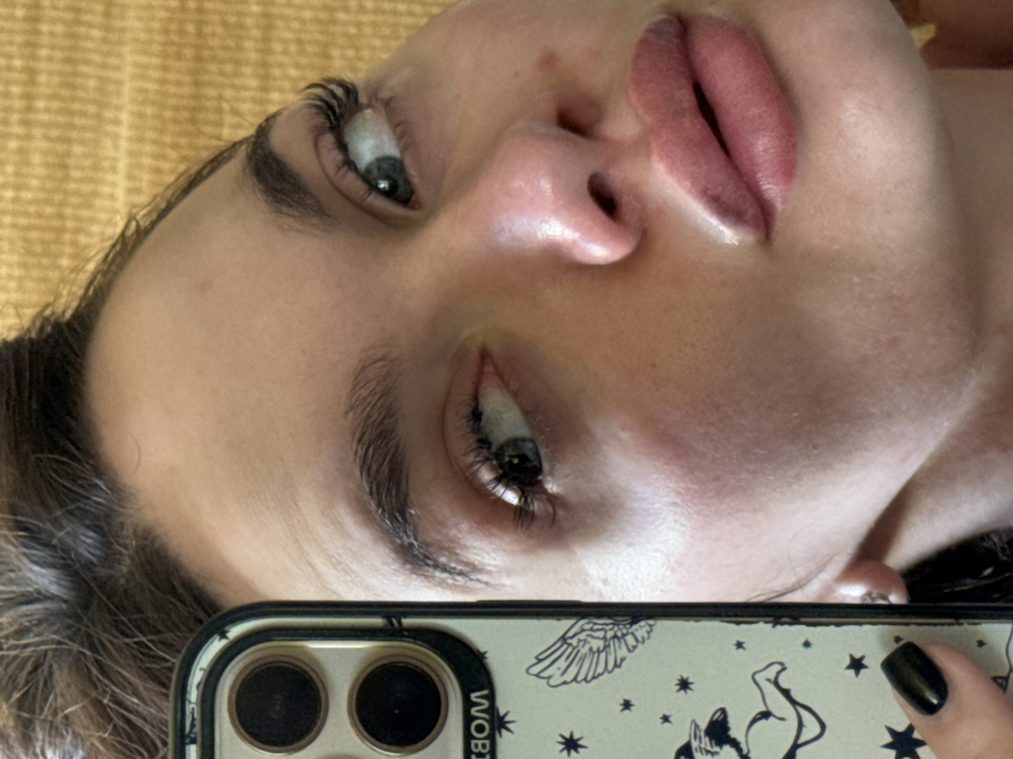 An angled close-up photograph shows a person's face from an upward-facing perspective. The person has light skin, green eyes, prominent lips, and dark eyebrows. The person's skin is shiny, possibly due to makeup or natural oils. The left side of their face is partially obscured by a phone. The phone case is patterned with white illustrations, including what appears to be an angel wing and other design elements, on a light background. A hand with a black polished fingernail is visible holding the phone. The background is a blurred yellow texture. The location is not discernible as there are no landmarks or other identifying features of Sofia, Bulgaria in the image.