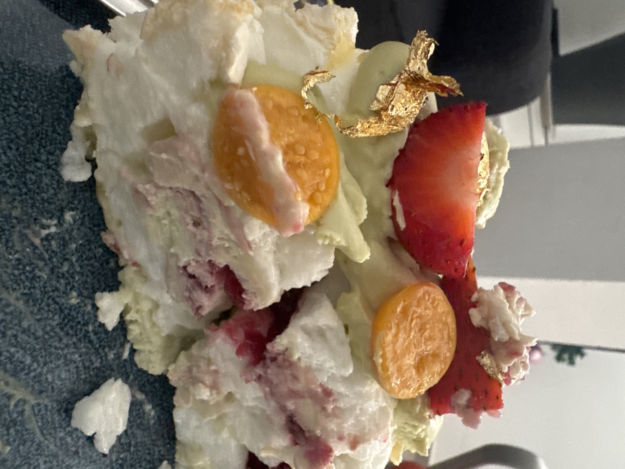 The image shows a close-up of a dessert, possibly Pavlova or a similar meringue-based confection, featuring a crushed meringue base topped with whipped cream, sliced strawberries, gooseberries, and edible gold leaf. The dessert appears to be on a plate. Given the prompt's request to include location in Ramat Gan, Israel, one can infer this image was taken within a food establishment in that city.