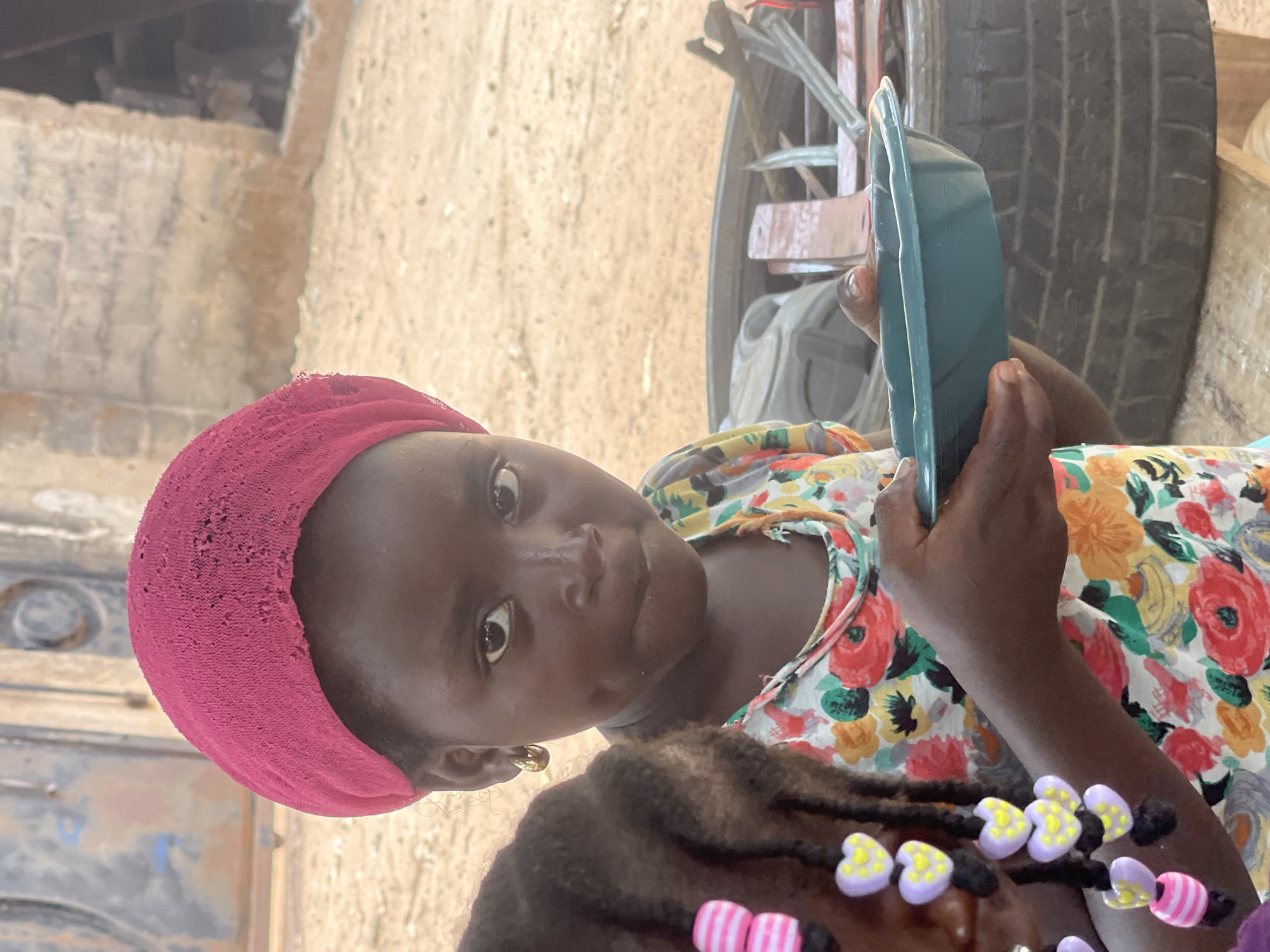 A close-up shot depicts a young girl with dark skin and a pink headscarf, adorned with hair braids featuring colorful beads. She gazes at the camera while holding a light green object in her hand. She is wearing a floral-patterned shirt. A tire is visible in the background, along with some building elements. The photo was taken outdoors, likely in Akwanga, Nigeria.
