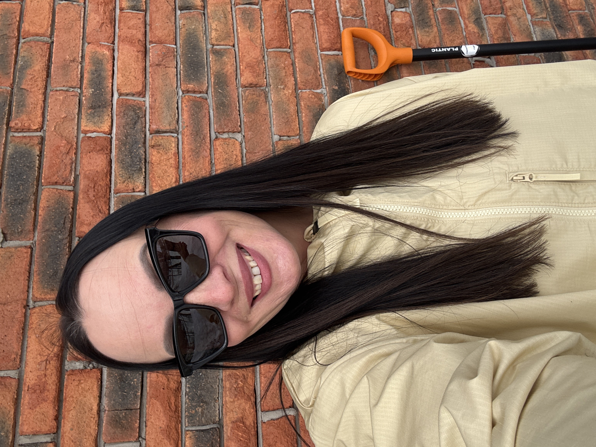 A selfie is taken outdoors on a brick patio in Chaikovsky, Russia. A woman with long dark hair, wearing sunglasses, lies on the brick surface. She is smiling. She is wearing a light yellow jacket. Next to her lies a black gardening tool with an orange handle.