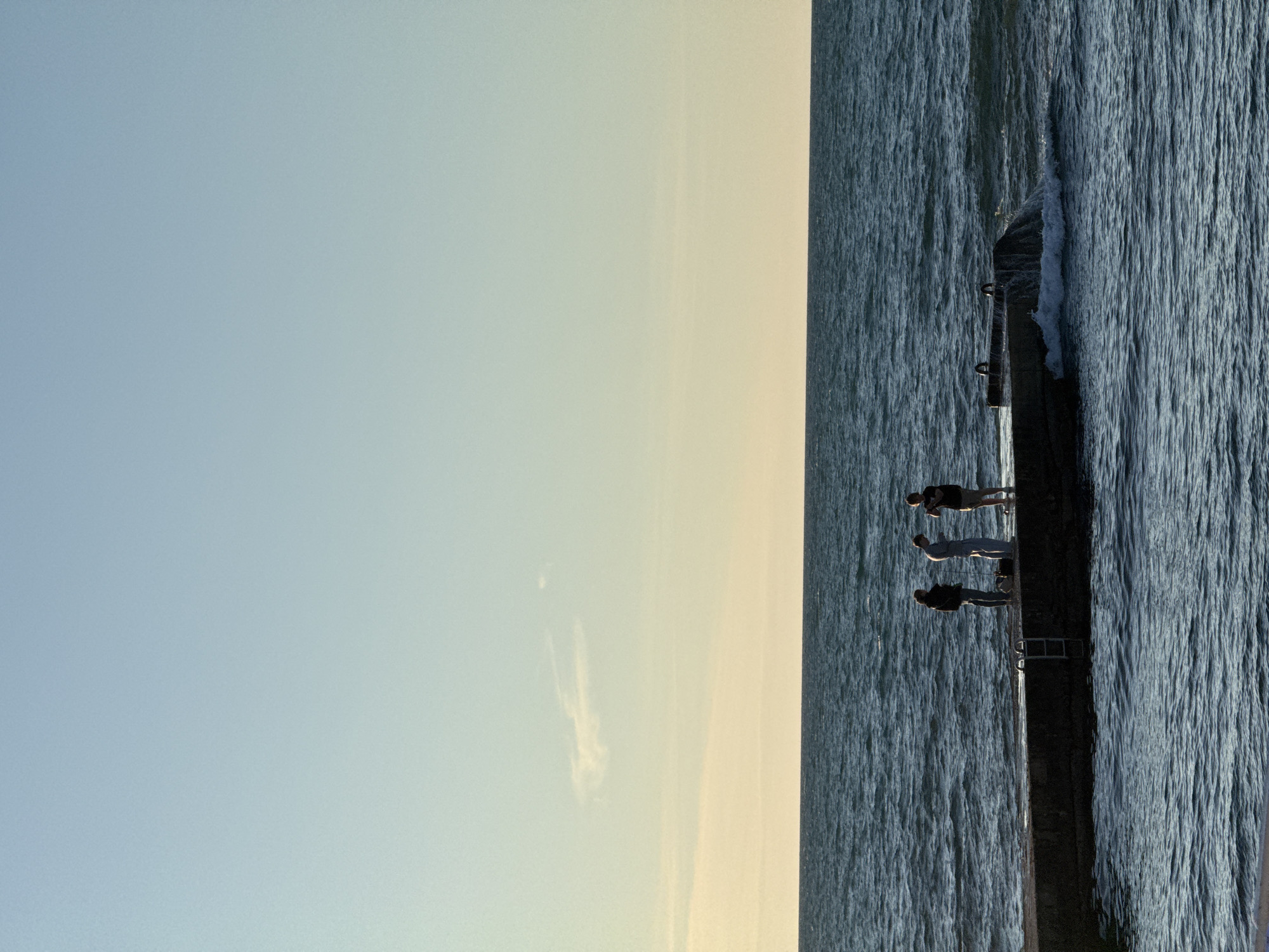 Three individuals stand on a concrete pier extending into the ocean. The sky is clear and exhibits a gradient of blue and pale yellow hues, suggesting either sunrise or sunset. The ocean is calm with small waves lapping against the pier. The individuals are silhouetted against the brighter sky. One person on the left appears to be holding a fishing rod, and another on the right is looking at a phone or small device. The pier has a metal ladder on its side, and there are some metal rings on the top surface. The overall scene is serene and captures a moment of outdoor activity by the water.