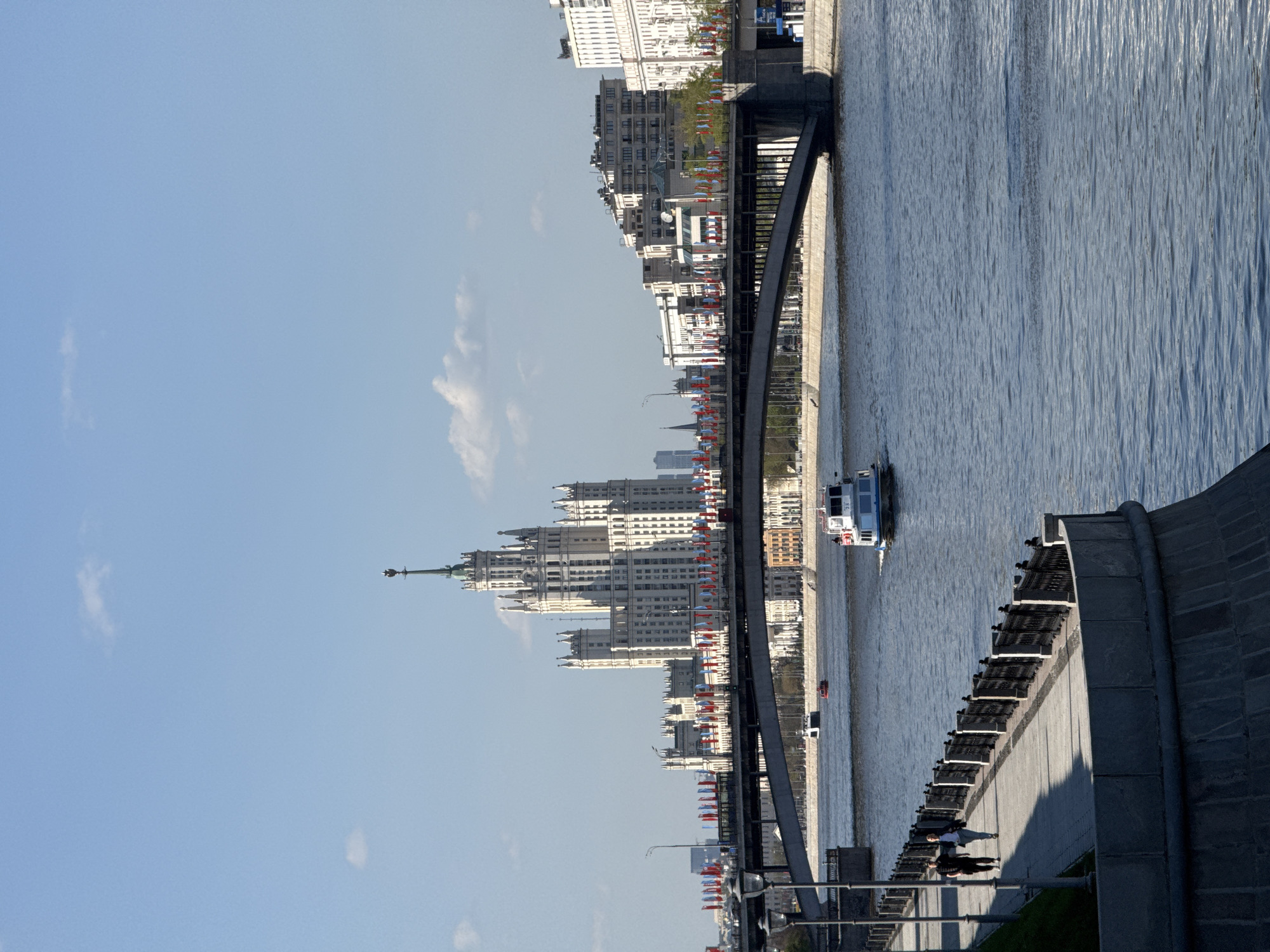 The image shows a view of the Kotelnicheskaya Embankment Building in Moscow, Russia, next to the Moskva River. The skyscraper is the dominant feature, with its distinctive Stalinist architecture. A bridge spans the river in the foreground, and a tour boat is traveling on the water. The sky is blue with scattered clouds. The embankment is visible with benches, and there are people sitting and walking. Decorations or flags are prominently displayed along the street and on the building.