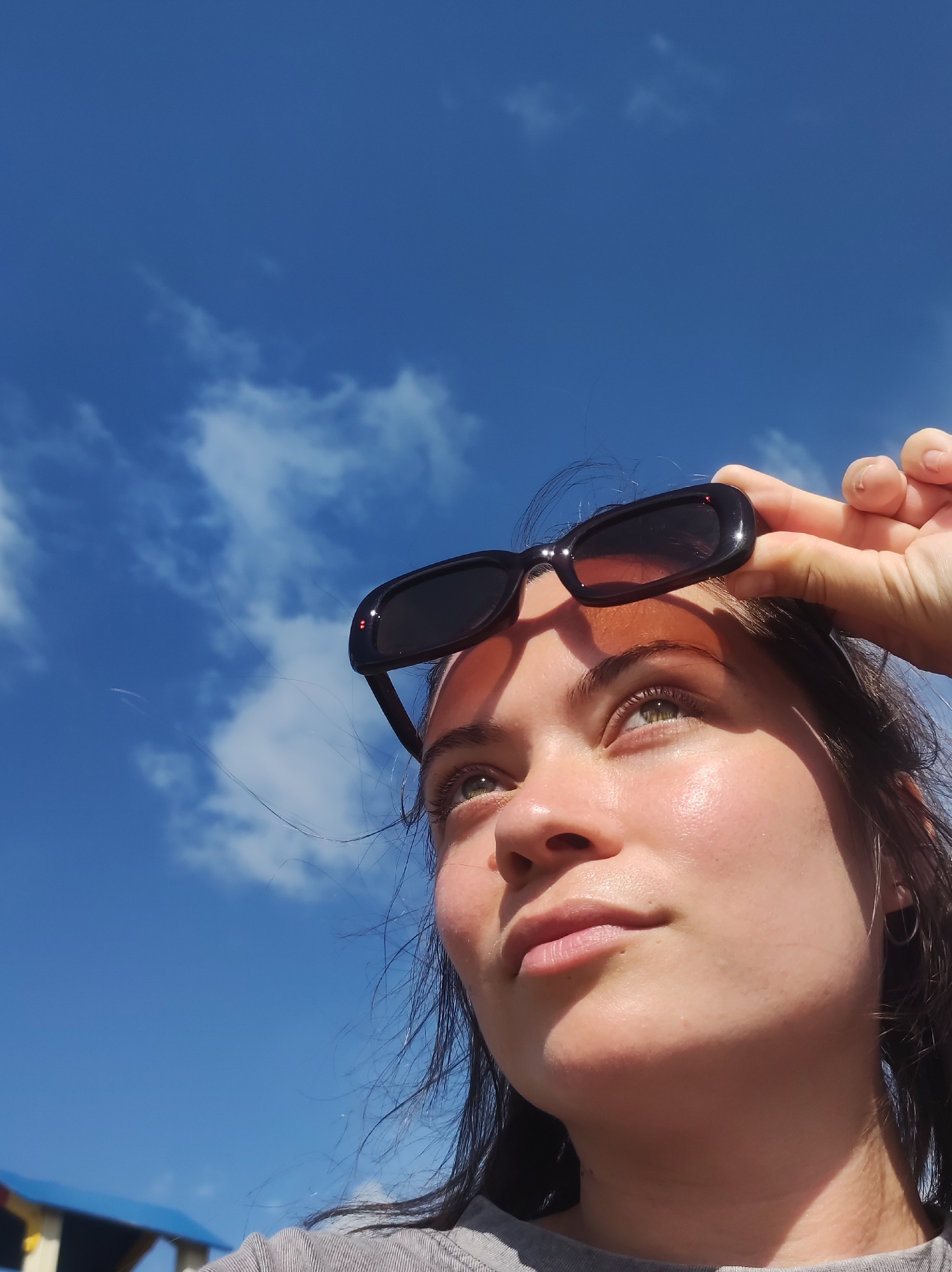 A young woman with light skin and green eyes is looking up at a bright blue sky with scattered white clouds. She is holding a pair of black sunglasses above her eyebrows with her right hand. The sun is casting a shadow on her face. She is wearing a grey t-shirt. In the background, the top of a playground structure is visible. The location is the city of Foky, Russia.