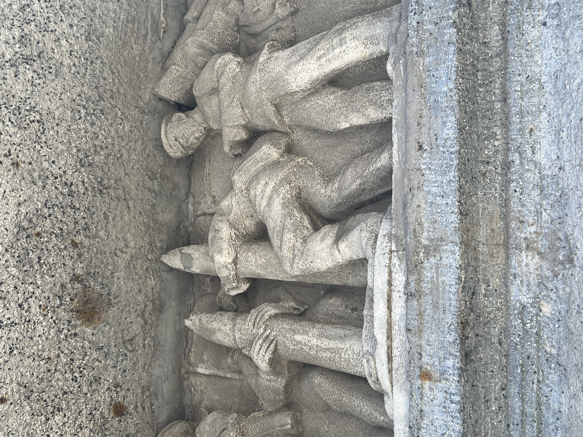 The image depicts a section of a stone bas-relief sculpture, likely on a building exterior, portraying human figures stacked in horizontal layers. The figures appear to be holding cylindrical objects, possibly weapons or tools. The style of the sculpture suggests a somber, possibly war-related theme. Given the location of Saint Petersburg, Russia, it is possible this is a section of the Memorial to the Heroic Defenders of Leningrad on Victory Square. The sculpture is aged and shows signs of weathering.