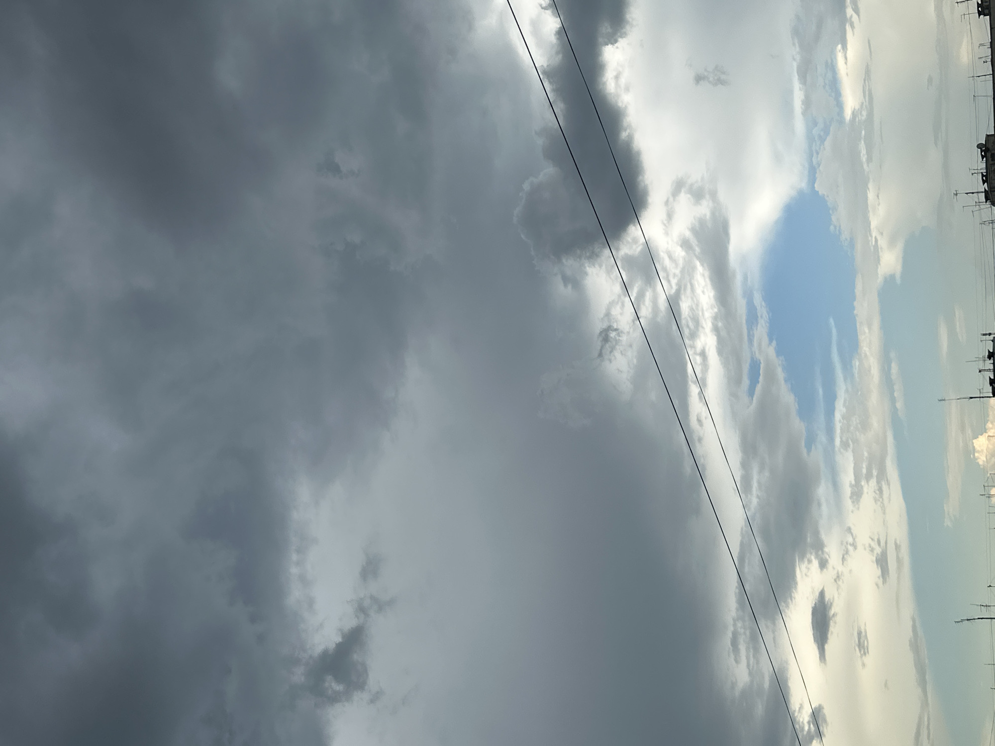 The image shows a dramatic sky filled with dark, heavy clouds, suggesting an impending storm. Two power lines stretch horizontally across the frame, intersecting the clouds. In the lower portion of the image, a patch of blue sky is visible, with lighter, scattered clouds below. The very bottom of the frame shows a blurry silhouette of buildings and antennas against the bright sky. The scene appears to be an urban environment looking upwards at the sky.