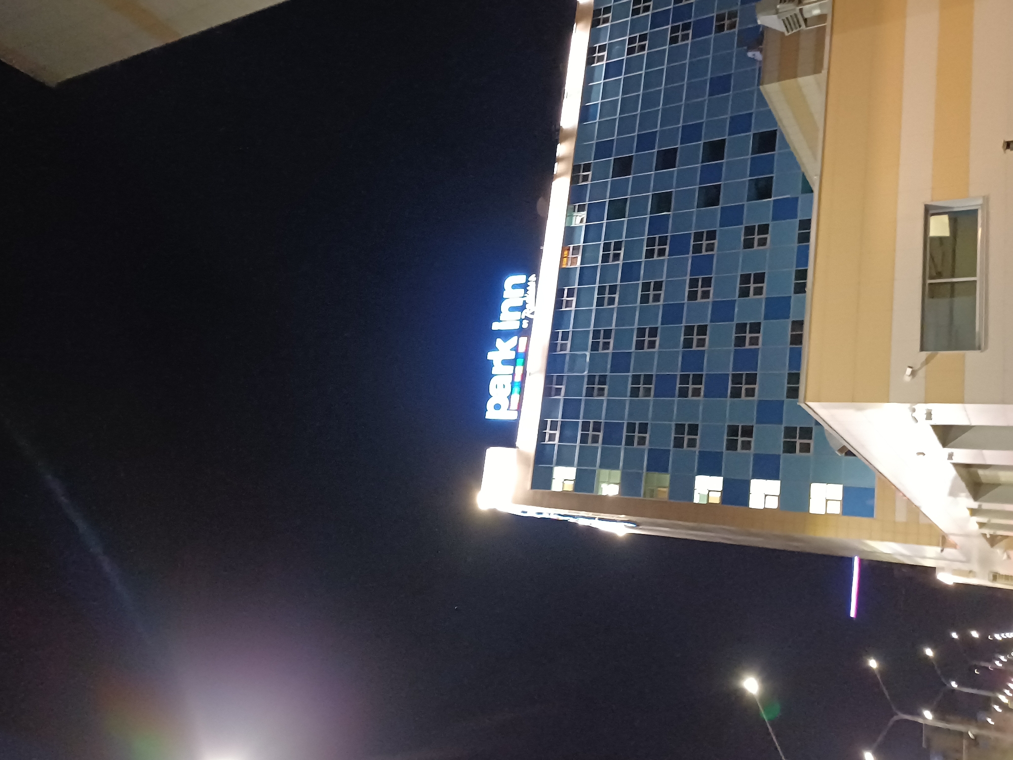 A wide shot captures the facade of a tall, modern hotel at night. The building's exterior is a grid of light blue and dark blue squares, punctuated by numerous windows, some of which are illuminated. At the top of the building, a large, bright neon sign reads "Park Inn" in blue letters, with a smaller "by Radisson" underneath. The sky above is a deep, dark blue. In the foreground, streetlights cast a glow on the scene, with one prominent streetlight on the left side of the image emitting a bright white light with a green lens flare. A portion of a lighter-colored building or structure is visible in the lower right corner. The image is slightly grainy, suggesting it was taken in low light conditions.