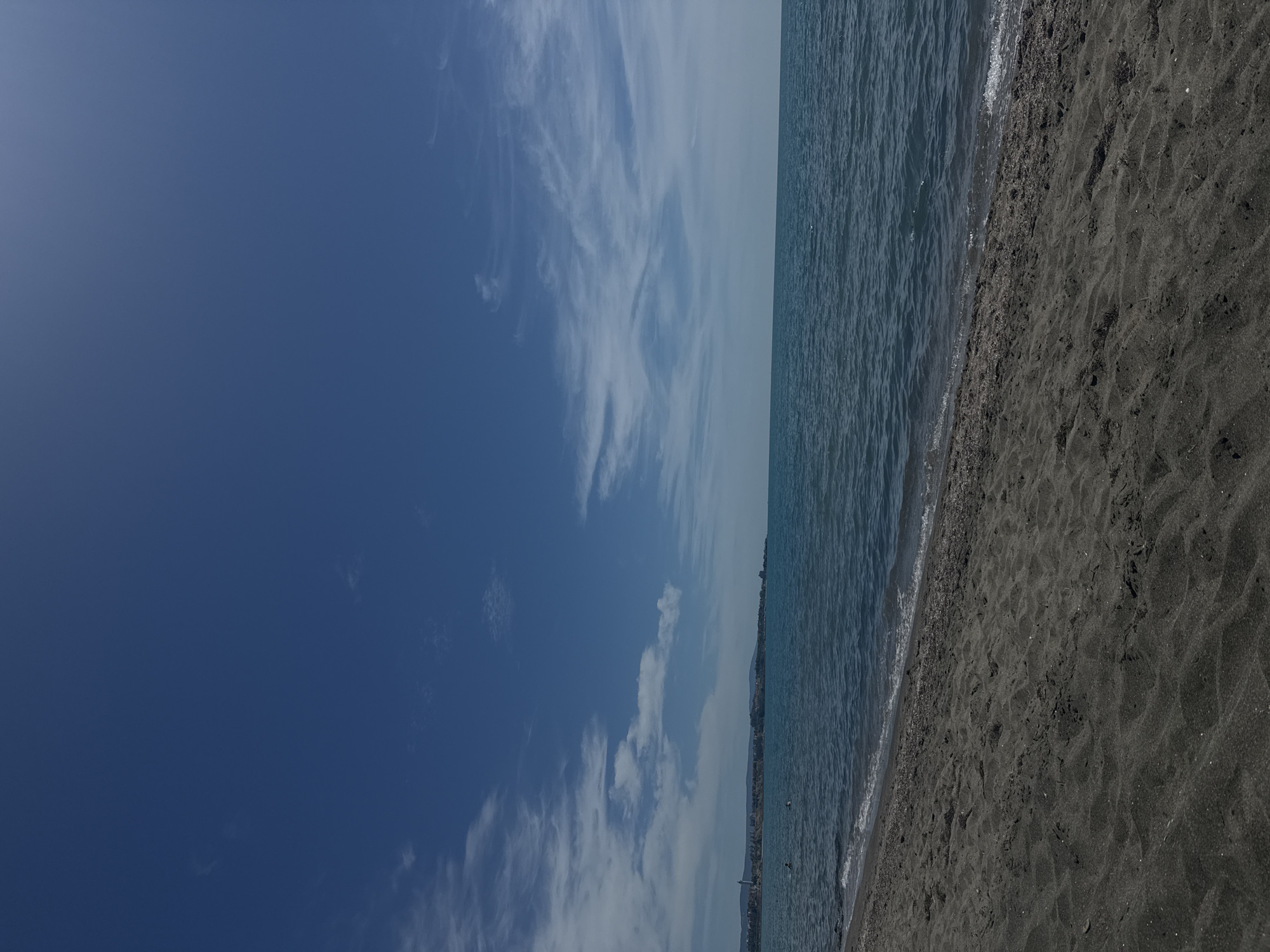 The image depicts a serene beach scene in Aheloy, Bulgaria, under a clear blue sky dotted with wispy clouds. The foreground is dominated by dark, coarse sand with visible ripples from wind or water. The dark sand transitions to a calm, deep blue sea that occupies the middle ground. The waterline shows gentle waves lapping at the shore. In the distance, along the horizon, a town or city is visible, characterized by buildings and some greenery, suggesting it is Aheloy itself. A faint silhouette of mountains can be seen behind the town. A few individuals are visible in the water, appearing to be swimming or wading. The overall impression is one of a tranquil day at the seaside.