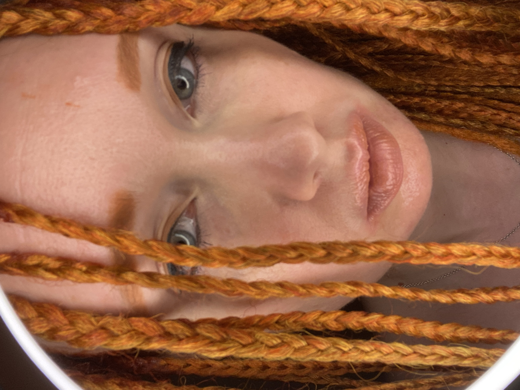A close-up, head-on, eye-level, medium shot captures the face of a Caucasian female with vibrant orange dreadlocks. Her eyes, a pale blue-grey, are framed by dark eyeliner and long lashes. Her lips are slightly parted and appear to be coated with a shimmering, peachy-toned lipstick. Some of her dreadlocks fall forward, partially obscuring her face. The background is dark and out of focus, with a bright, circular light source visible on the left side of the frame, likely a ring light. The image is shot indoors, with no specific location in Sochi, Russia, identifiable.