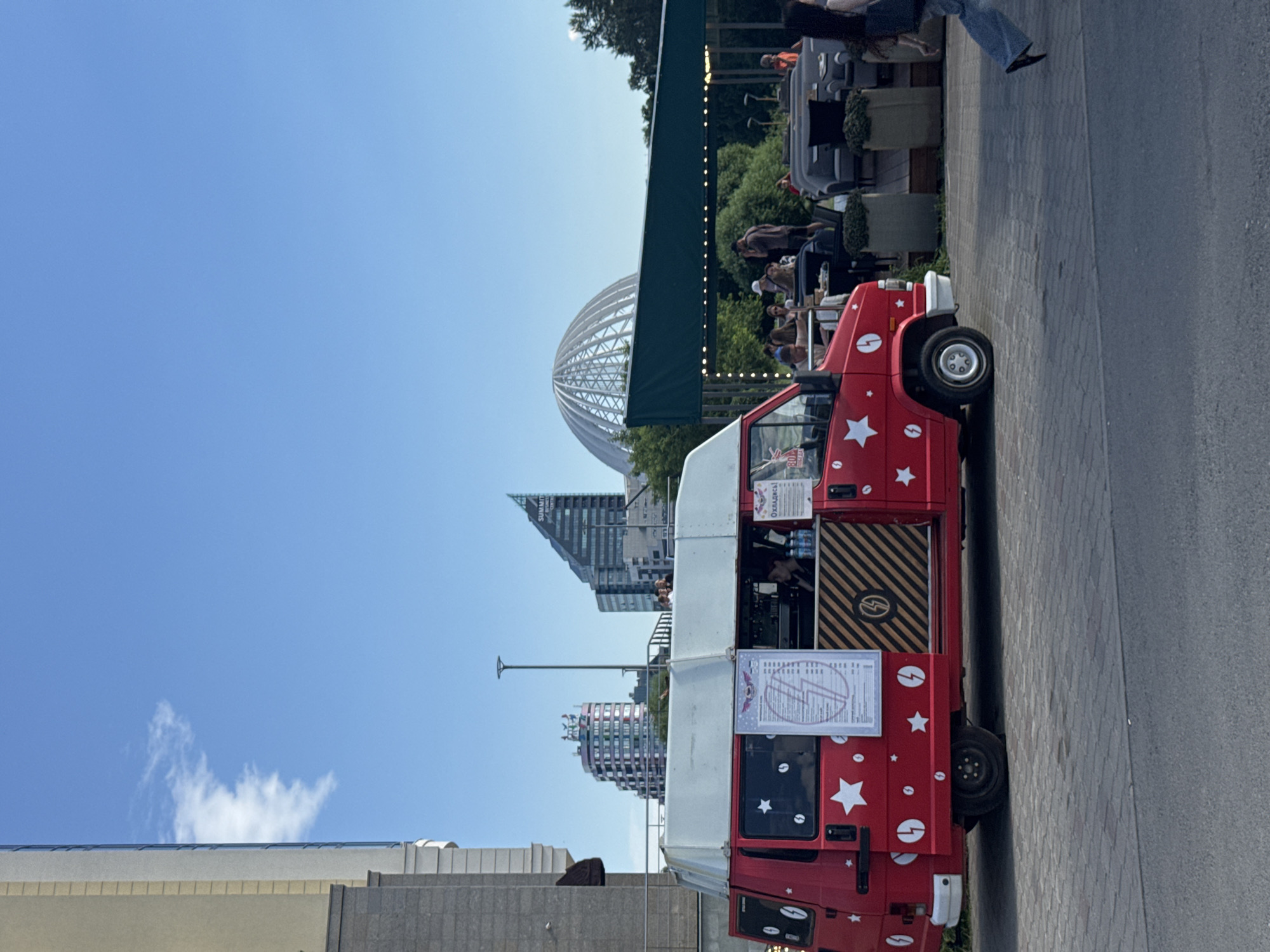 This image depicts a vibrant street scene in Yekaterinburg, Russia, likely in an urban park or pedestrianized area. In the foreground, a red food truck, decorated with white stars and lightning bolt symbols, is parked on a paved walkway. The truck appears to be operational, with a menu displayed and a person visible inside, suggesting it is serving customers. Behind the food truck, an outdoor seating area with tables and chairs is set up under a green canopy, with people gathered, enjoying food and drinks. In the background, modern city architecture is visible, including several buildings with distinctive designs. Notably, a large, geodesic dome structure stands out against the clear blue sky, a landmark that could help pinpoint the exact location within Yekaterinburg, possibly near the Yekaterinburg-City complex or the Yeltsin Center. The overall atmosphere is lively and social, indicating a pleasant day in the city.