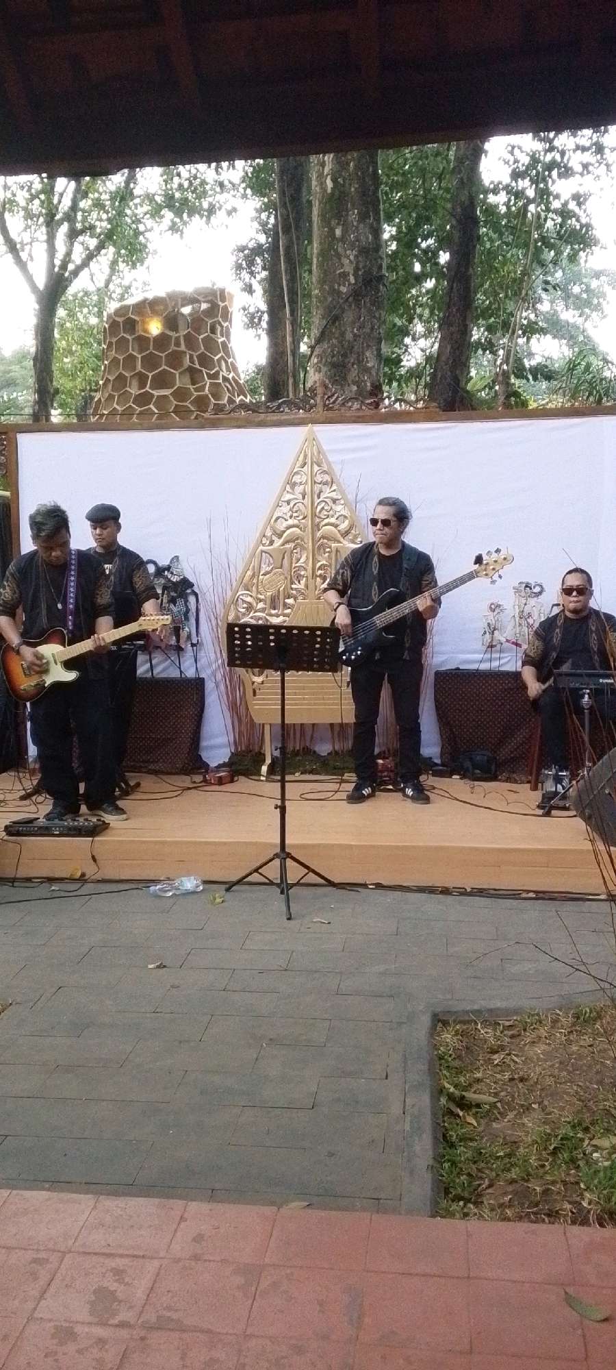 The image depicts an outdoor stage setup for a musical performance, likely in Central Jakarta, Indonesia, given the cultural elements present. Four individuals are visible on the stage, each engaged in playing musical instruments. The central backdrop features a large, ornate golden Javanese wayang (shadow puppet) silhouette, flanked by white fabric panels.
On the left, a person with dark hair and wearing a black patterned shirt and dark pants is playing a light-colored electric guitar. To their right, a second individual, also in dark clothing and wearing a flat cap, stands behind the first guitarist, appearing to be a backup musician.
In the center-right, a person with dark hair pulled back, wearing sunglasses and a black patterned shirt with dark pants, plays a black electric bass guitar. To their right, a fourth individual, also wearing sunglasses and a similar black patterned shirt with dark pants, is seated and appears to be playing a drum set, though only the top of the drum and cymbals are visible.
In front of the stage, a black music stand with sheet music is positioned. The stage itself is elevated and appears to be made of wood or a similar material. The surrounding environment suggests an outdoor setting, possibly a garden or park, with trees and foliage visible in the background. A distinctive, honeycomb-patterned structure, resembling a decorative chimney or sculpture, is visible behind the stage to the left. The foreground shows a paved walkway made of grey and red tiles. A discarded plastic water bottle lies on the ground in front of the stage. The overall impression is that of a live musical event taking place in a culturally significant location.