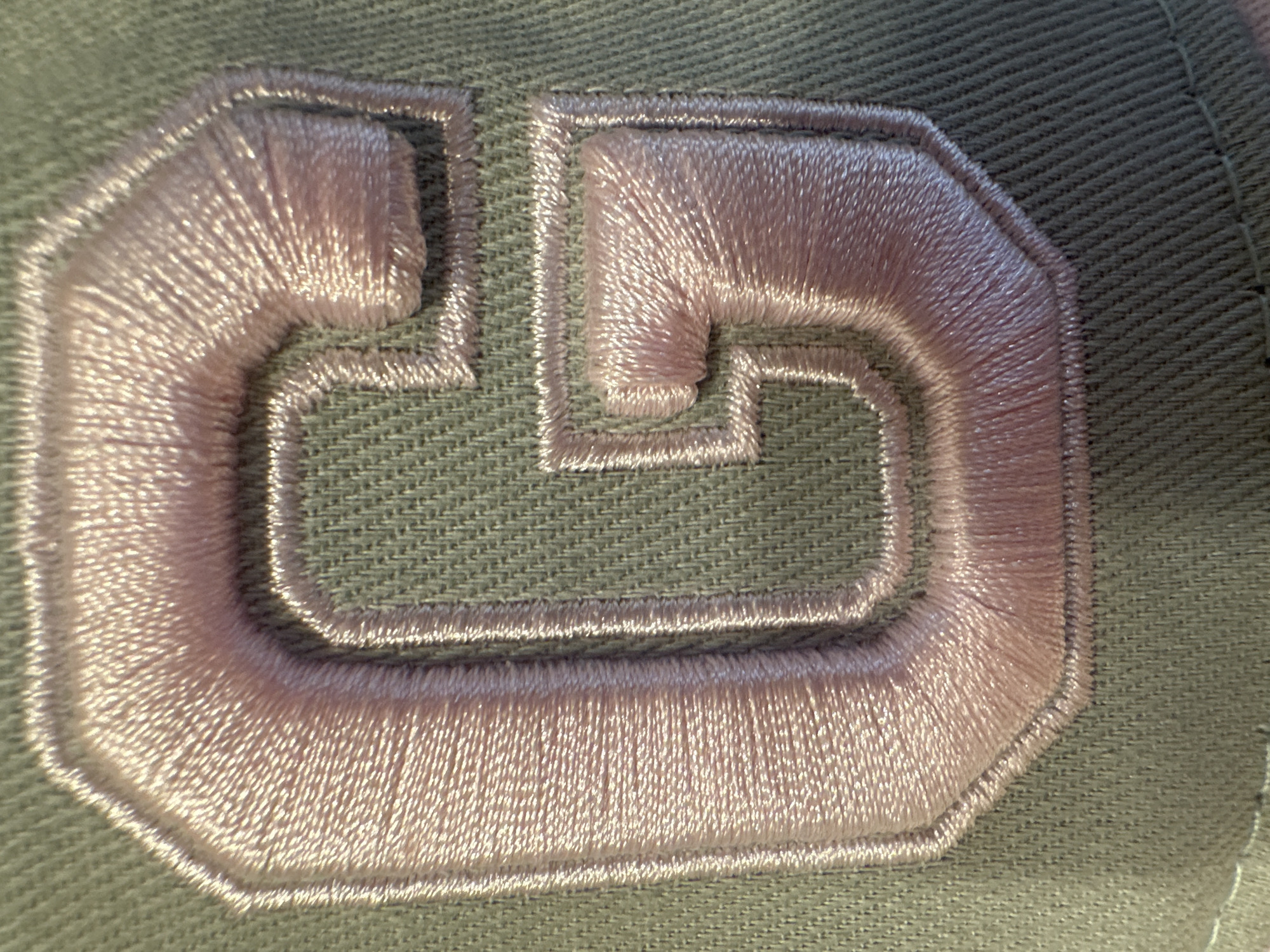 The image is a close-up, macro shot of an embroidered letter "G" on a textured, olive-green fabric, likely a cap or piece of clothing. The letter is outlined with a thinner, lighter colored thread, creating a subtle border. The main body of the "G" is filled with a shiny, light pink embroidery thread that reflects light, giving it a lustrous appearance. The stitching is dense and runs in a way that follows the curves and lines of the letter, creating a smooth and raised effect. The fabric underneath has a visible weave pattern, adding to the texture of the image. There is no text visible in the image. The lighting suggests it is daytime, with light casting highlights and shadows on the embroidery, emphasizing its three-dimensional quality. The setting appears to be indoors or a controlled environment, given the focus on the object.