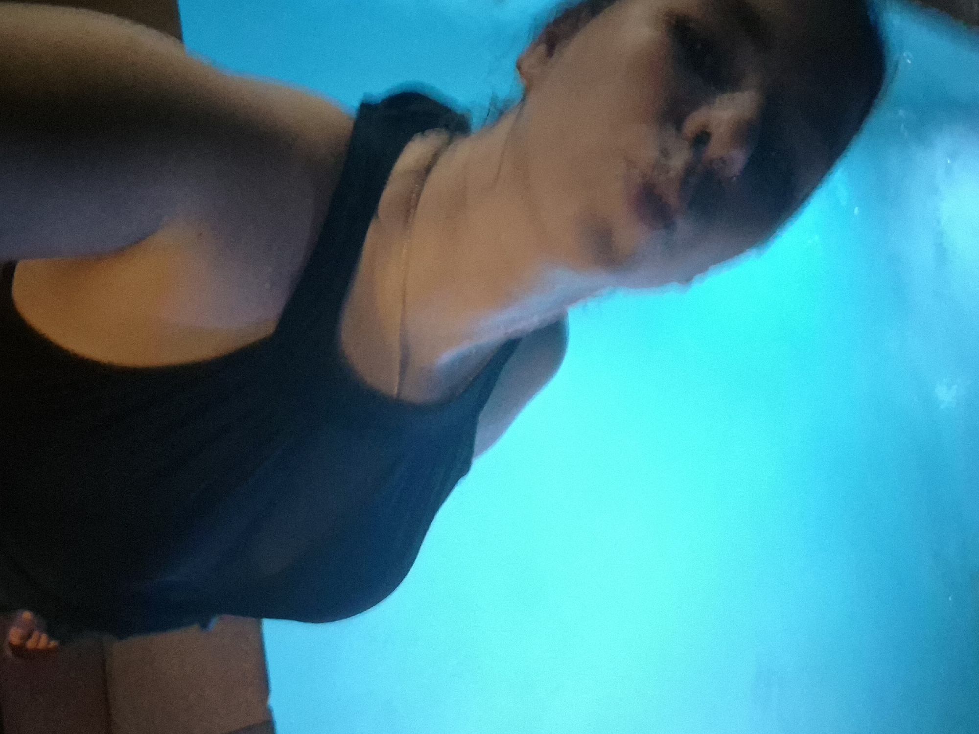 A woman wearing a black tank top is positioned in the upper left of the frame. Her face, partially blurred, occupies the upper right quadrant. The background is a vibrant turquoise color, possibly indicating water. A fragment of a tiled surface is visible in the lower left corner. There is insufficient information to ascertain the location in Chaikovsky, Russia.