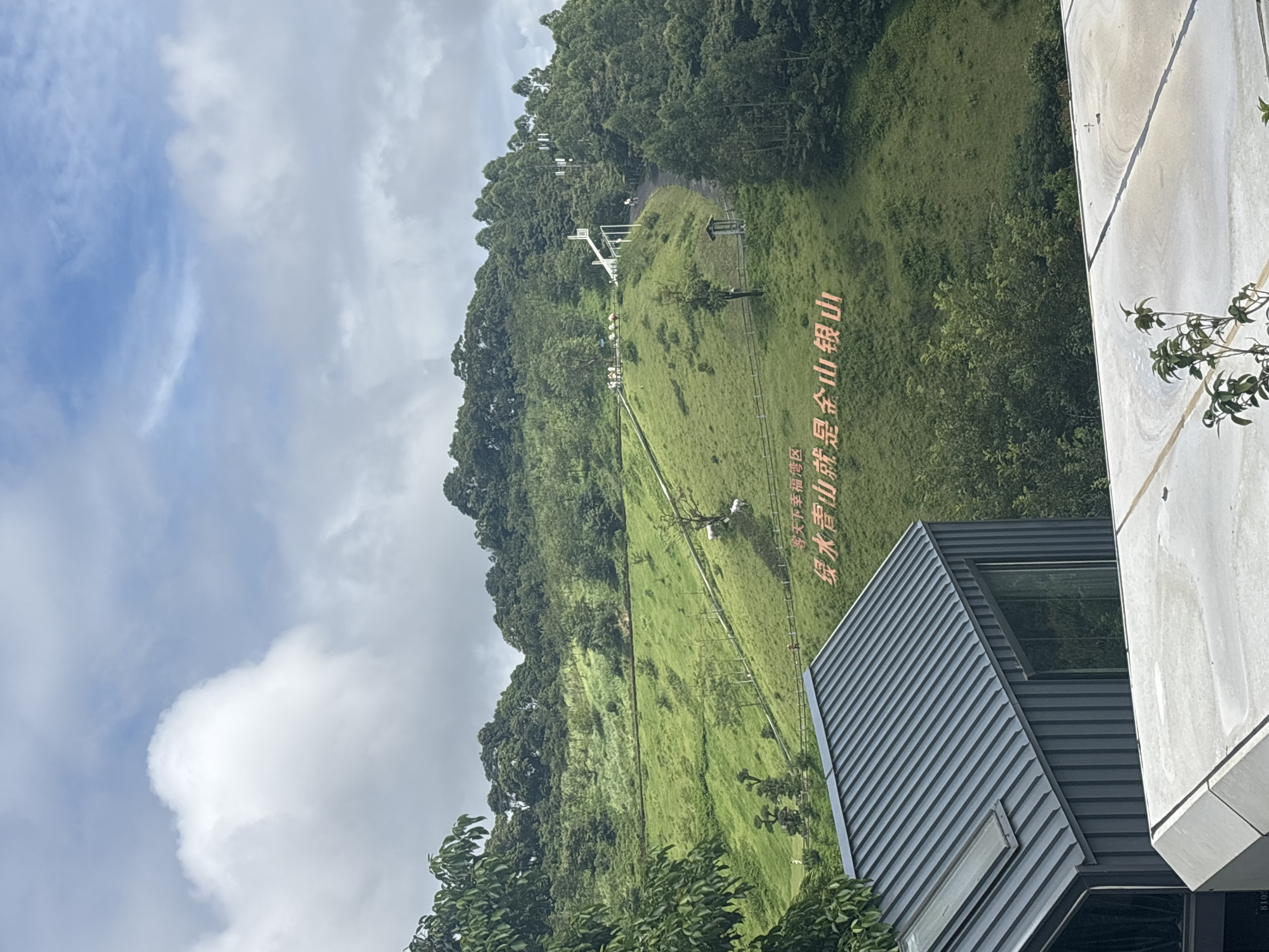 This image depicts a scenic view of a green hillside with trees and white structures against a cloudy sky. In the foreground, a modern building with a corrugated metal roof is visible. The hillside is covered in lush grass and dotted with trees. Red characters are inscribed on the hillside, reading "碧水青山就是金山银山" which translates to "Clear waters and green mountains are gold and silver mountains." This phrase is a well-known environmental slogan in China. In the distance, a white structure, possibly a slide or observation deck, is situated on the hilltop. Several people can be seen walking on pathways and near the white structure. The scene appears to be a tourist attraction or a natural park. The phrase on the hillside suggests that this location is associated with environmental conservation and natural beauty. Given the phrase's prominence and its association with ecological protection campaigns, it is likely that this is a location in Guangzhou City, China, that promotes eco-tourism or environmental awareness.