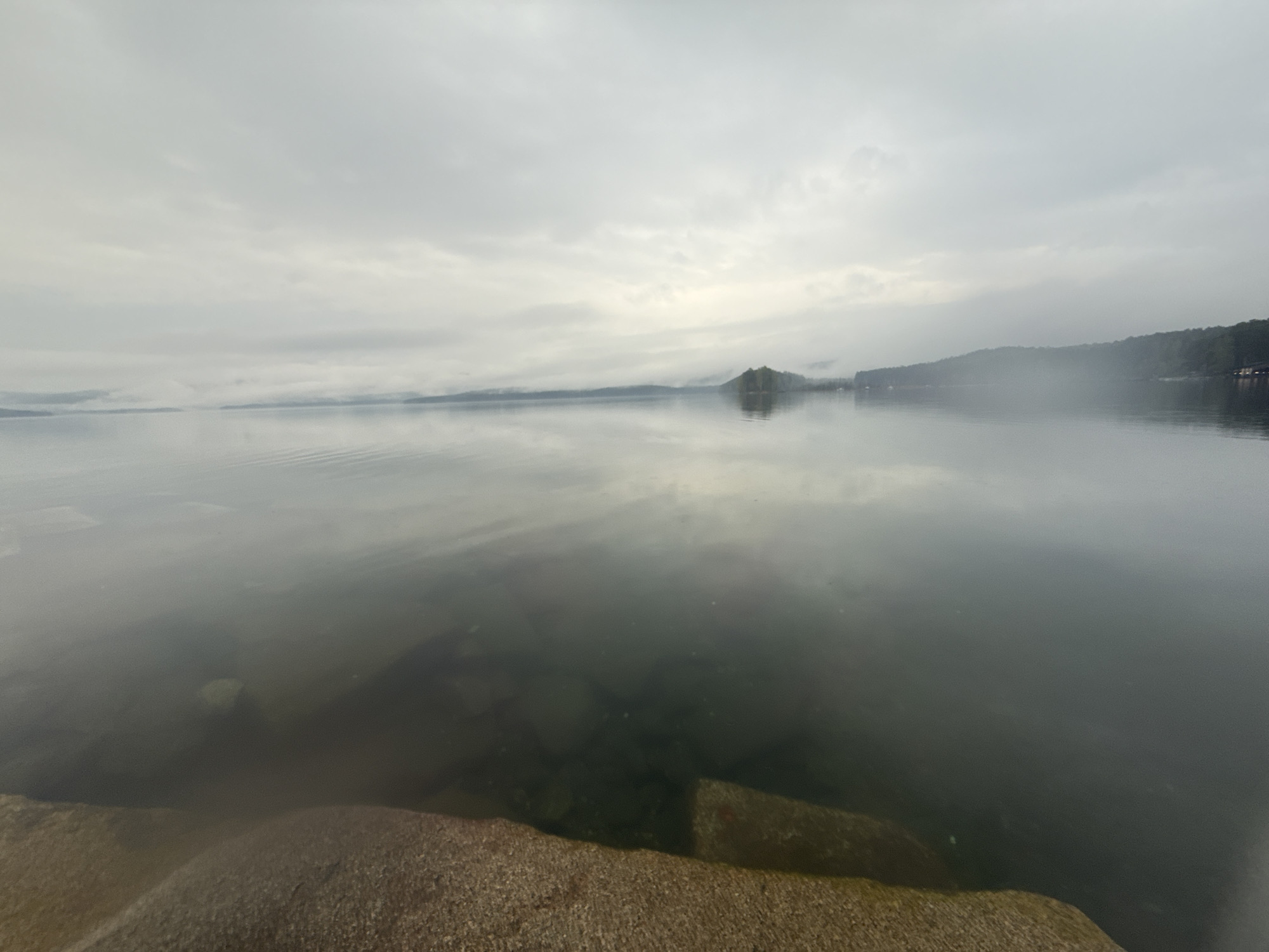 The image shows a body of water under a cloudy sky, likely a lake or bay. The water is calm with reflections of the sky and distant land. In the foreground, there are large rocks partially submerged in the water. The distant land appears to be forested hills or mountains, and there's a small island-like feature in the middle distance. The overcast lighting creates a muted, gray color palette. There is no definitive evidence to confirm that this image is of Zolotoi Plyazh, Russia.