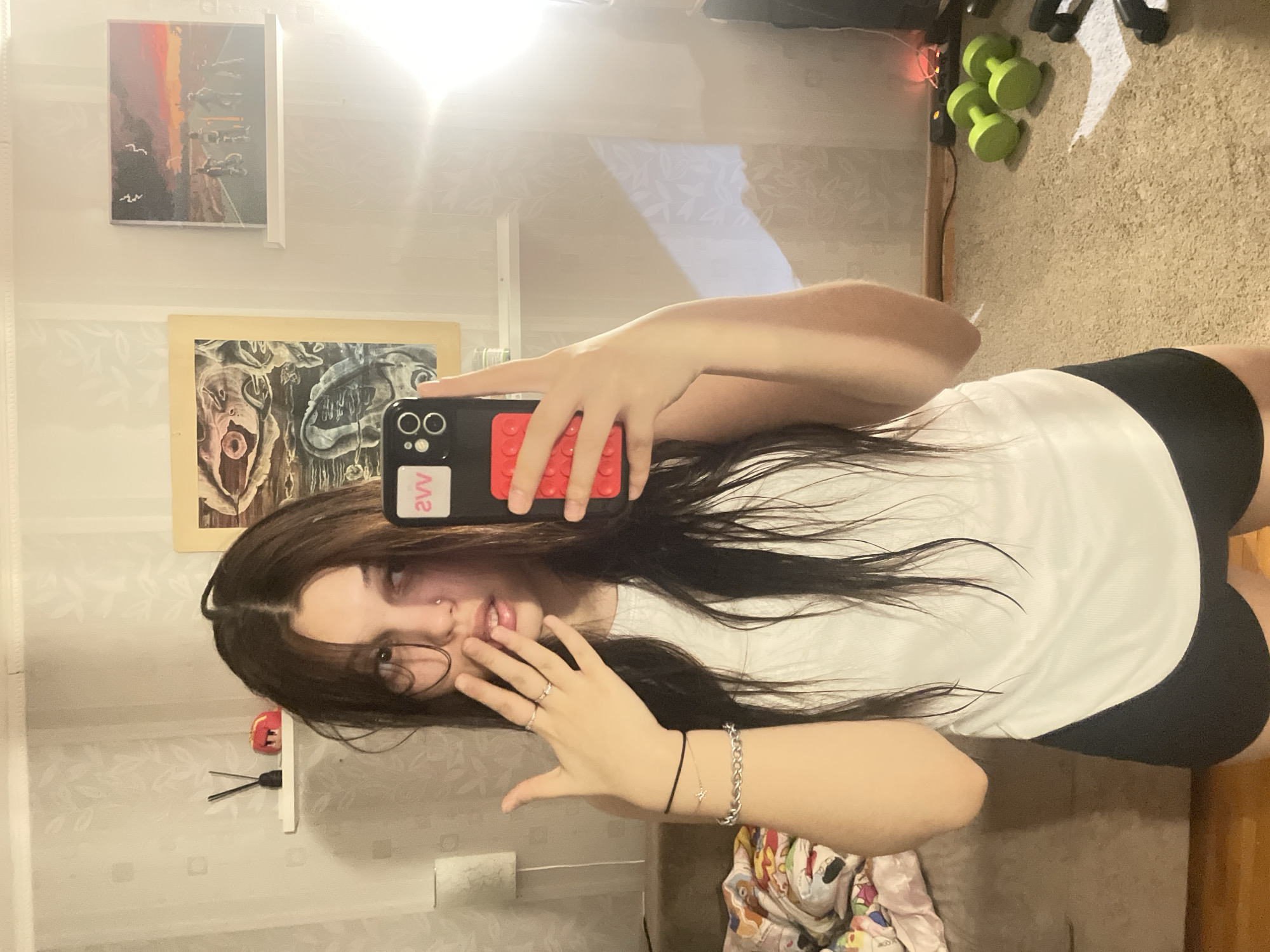 This image captures a young person taking a mirror selfie indoors in Saint Petersburg, Russia. The individual has long, dark hair with a middle parting, and a subtle smile. They are wearing a white t-shirt and black shorts. Visible accessories include a nose piercing, multiple rings on their left hand, and two bracelets (one chain-link, one delicate string) on their left wrist.
They are holding a black smartphone, which appears to be an iPhone with a triple camera setup, in a red, textured case resembling Lego bricks. A white sticker with black text, displaying "2VV" (reversed in the reflection), is affixed to the back of the phone.
The background, reflected in the mirror, shows a carpeted floor on the right side of the frame, where three green dumbbells and some string lights are visible. The walls are adorned with subtly patterned wallpaper. Two paintings are hanging: one abstract artwork with warm hues and silhouetted birds, and another, more surreal piece with darker, organic shapes. A small white shelf holding a red object and black wires can also be seen. The room is brightly illuminated by an overhead light source, causing a prominent glare in the upper part of the mirror. The scene depicts a casual, personal moment.