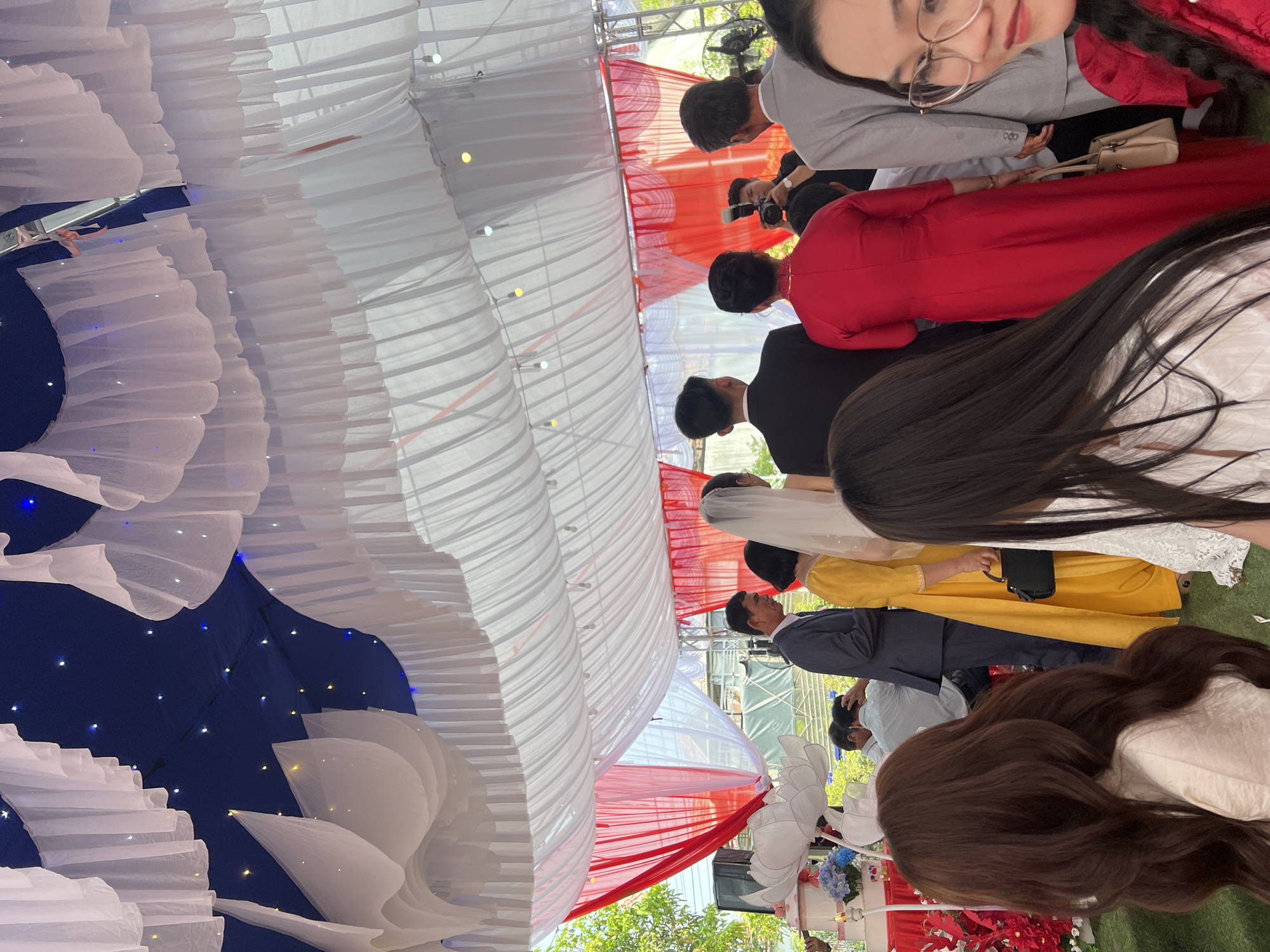 This image captures an outdoor celebration, likely a wedding, taking place under an elaborately decorated canopy in Tay Son Commune, Vietnam. The scene is filled with guests, many dressed in formal attire.
The canopy ceiling is a prominent feature, adorned with cascading white ruffled fabric. Sections of dark blue fabric embedded with numerous small fairy lights create a star-like effect, while panels of red and white striped fabric add to the festive decor. Beneath this, a diverse group of people gathers. In the immediate foreground, a woman with long dark hair and glasses smiles faintly towards the camera. She wears a grey top, and next to her, another person in a traditional red Vietnamese Ao Dai is visible from the back, holding a beige handbag.
Further back, a bride in a white wedding gown and veil is seen from behind, accompanied by a guest in a bright yellow dress. Several men in suits, including a presumed groom in a black suit and a guest in a grey shirt holding a camera, are also present. The event appears to be held on a green surface, possibly turf or artificial grass. Natural daylight indicates it's daytime, while the presence of a fan suggests warm weather. Some green foliage is visible outside the canopy, and artificial red and blue floral arrangements adorn a stand or table. The overall atmosphere is one of celebration and joyous gathering. No discernible text is visible in the image.