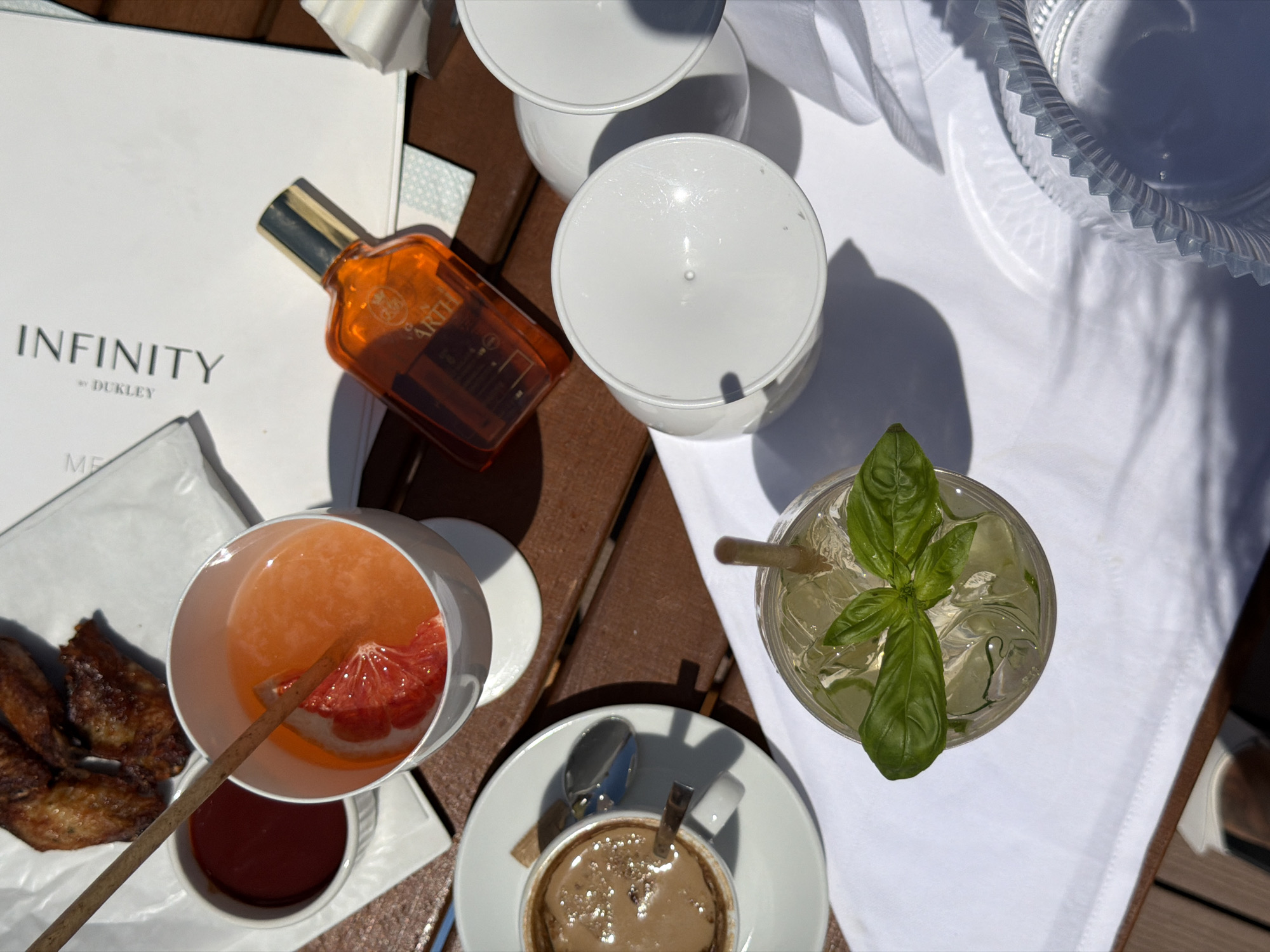 An overhead shot depicts a wooden table with a white cloth laid out. On the cloth, several items are arranged, including a glass of orange liquid with a slice of grapefruit and a wooden stirrer, a cup of coffee with a spoon, a glass of clear liquid with ice and a basil sprig, and two empty white cups. A small white dish of red sauce sits next to a plate of three fried chicken wings. A small orange bottle with a gold cap is placed on a folded white document. The scene appears to be in Bečići, Montenegro.