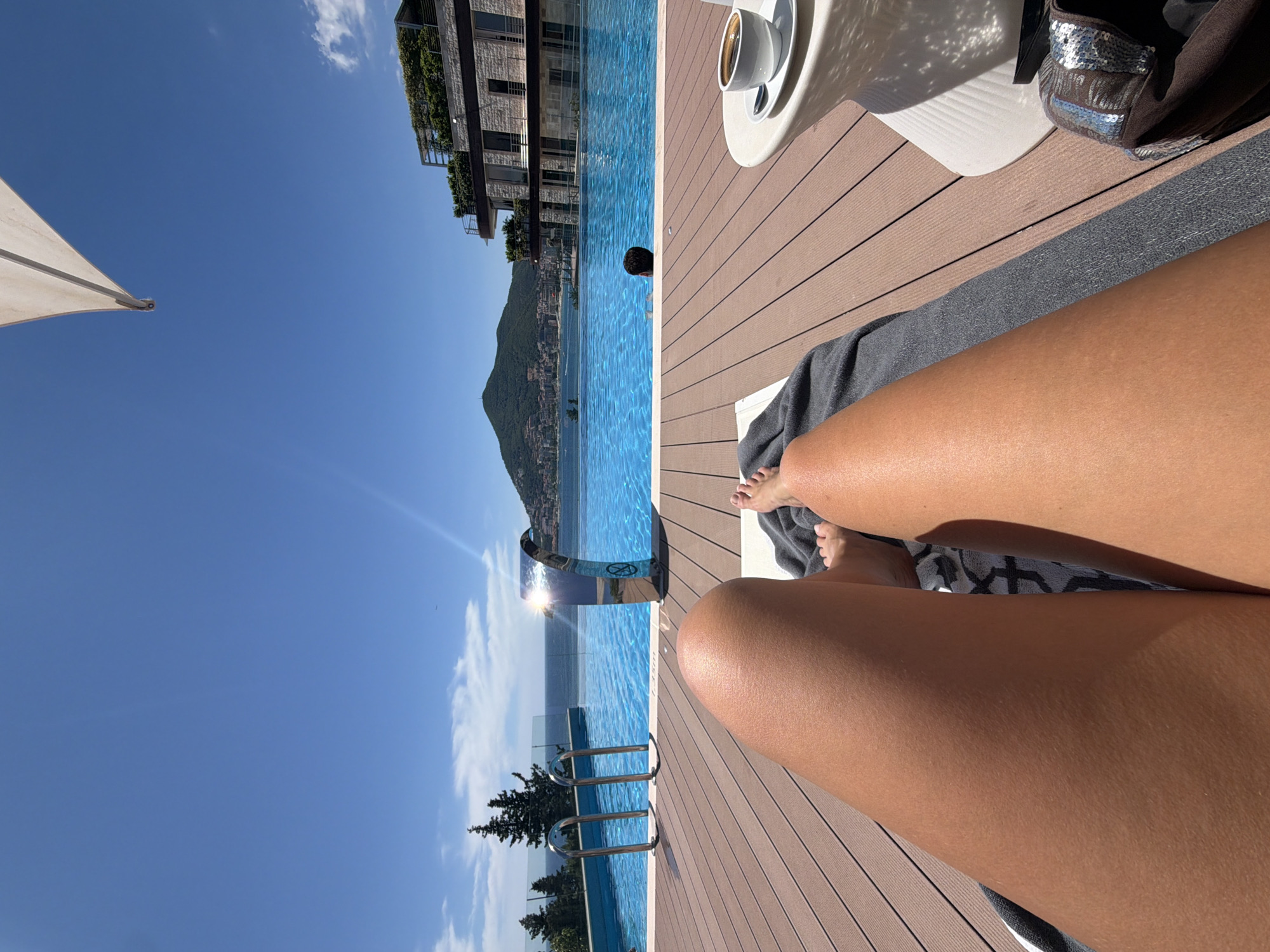 The image depicts a first-person perspective of someone relaxing on a lounge chair by an infinity pool overlooking the Adriatic Sea. The pool is a vibrant blue and has a waterfall feature. To the right, a white side table holds a cup of coffee. In the background, a modern building with balconies and greenery is visible, along with a hilly coastline characteristic of Bečići, Montenegro. The sky is clear blue with a few wispy clouds. A person's legs are prominently in the foreground, with their feet visible on the edge of the lounge chair. Another person is partially submerged in the pool in the mid-ground. The wooden deck surrounding the pool is a dominant feature of the foreground.