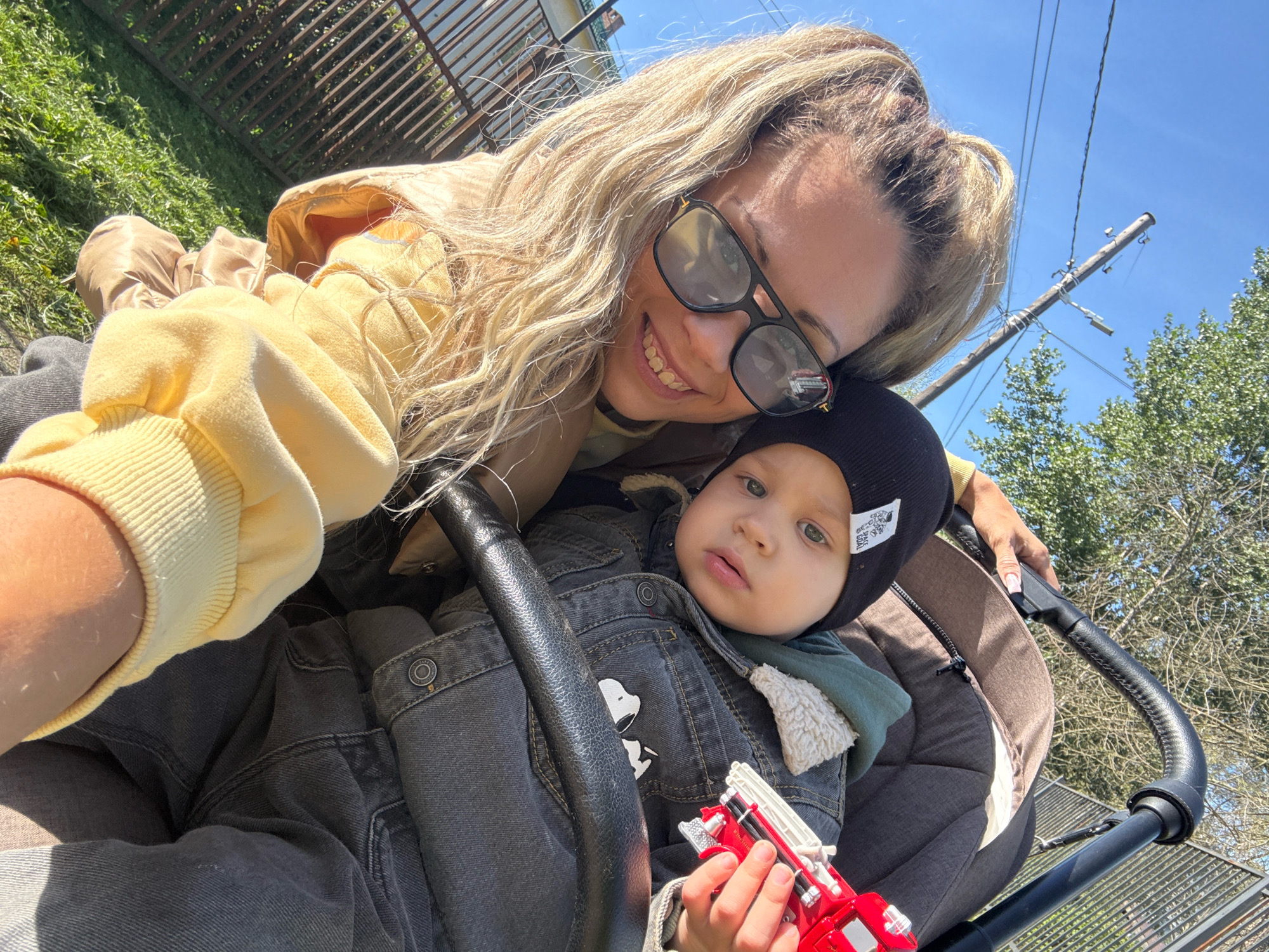 In Rusanovo, Russia, a woman with blonde hair and sunglasses takes a selfie with a young child in a stroller. The child, wearing a black beanie and a denim jacket with a Snoopy patch, holds a toy fire truck. The background features a wooden fence, green grass, trees, and a clear blue sky. The scene appears to be outdoors during the daytime, possibly in a park or residential area.
