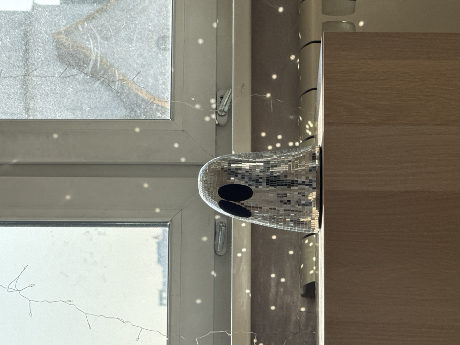 The image depicts an indoor scene in Moscow, Russia, looking outward through a window. A stylized ghost-shaped object, approximately 20-30 cm in height, constructed from small, reflective mirror tiles akin to a disco ball, is positioned centrally on a light-toned wooden surface in the foreground. This object features two prominent, oval-shaped black cutouts representing eyes.
Behind the object is a white-framed window, divided into multiple panes. Through the left window pane, a hazy, bright sky and indistinct silhouettes of distant urban buildings are visible. The right pane offers a blurred view of an exterior structure or ground. Thin, white string lights are draped across the window frames and extend partially onto the glass. Numerous small, bright, circular light reflections are scattered across the window panes, the white window sill, and the wall below, indicating ambient light interaction with the mirrored object and possibly the string lights. A white, horizontally finned radiator is partially visible below the window sill on the right. No individuals or active interactions are present within the frame.