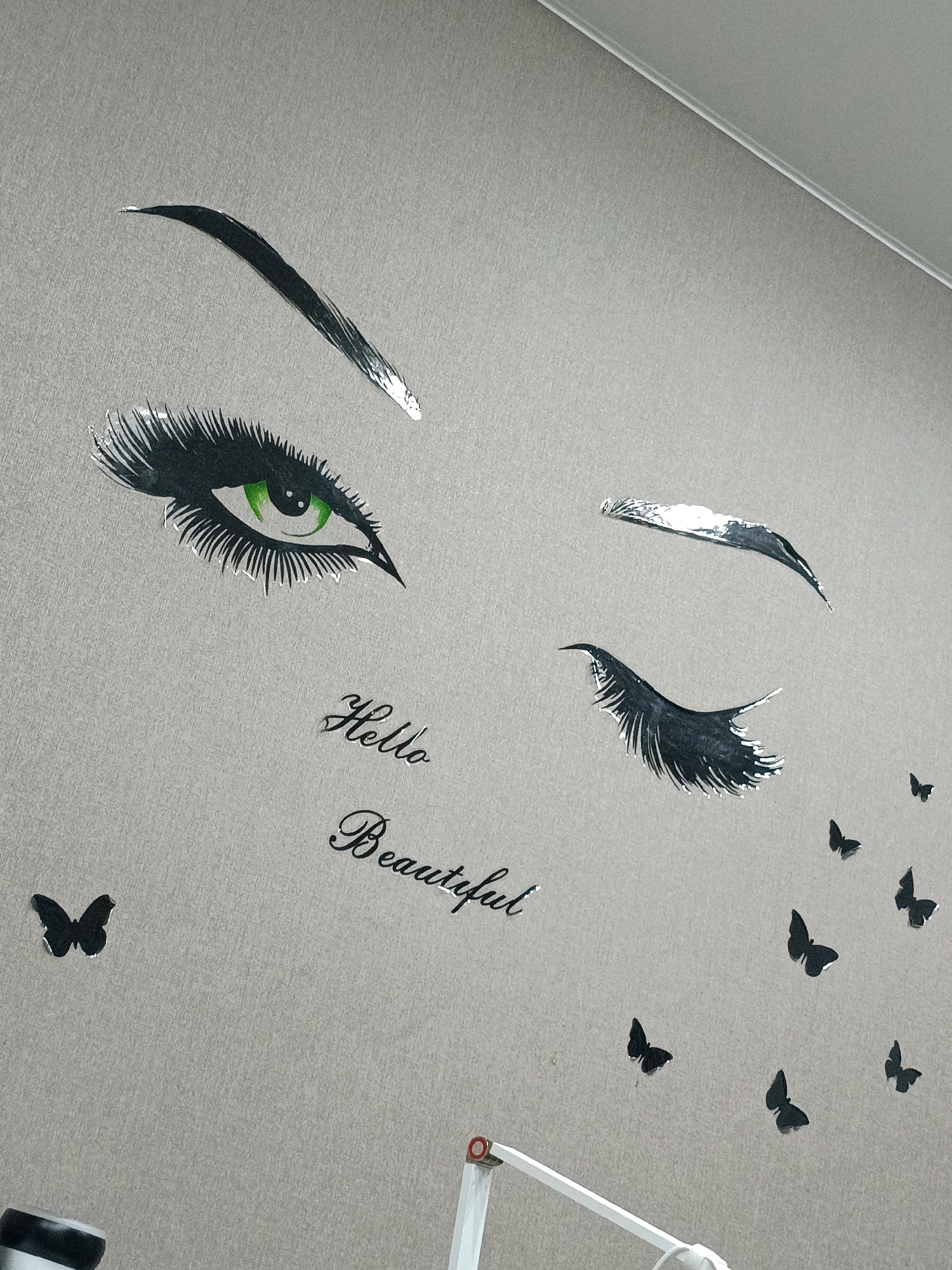 The image shows a wall decorated with decals of a woman's eye and eyelashes, the words "Hello Beautiful," and several butterflies. The eye is depicted with a green iris and long, dark eyelashes. The decals are silver and black. The wall itself is a textured light gray. There is a portion of a white lamp and its stand visible in the bottom right corner of the image. The location of this scene within the city of Asbest, Russia, is not ascertainable from the provided image.