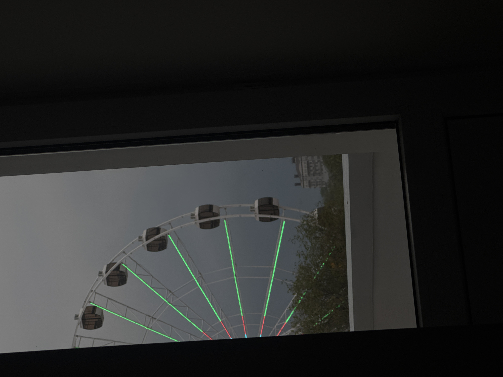 The image depicts a partial view of a Ferris wheel through a window. The Ferris wheel is white with dark passenger cars. Green and red lights are visible on the spokes. A tree and a building can be seen in the background. The location is Yevpatoriya, Ukraine.