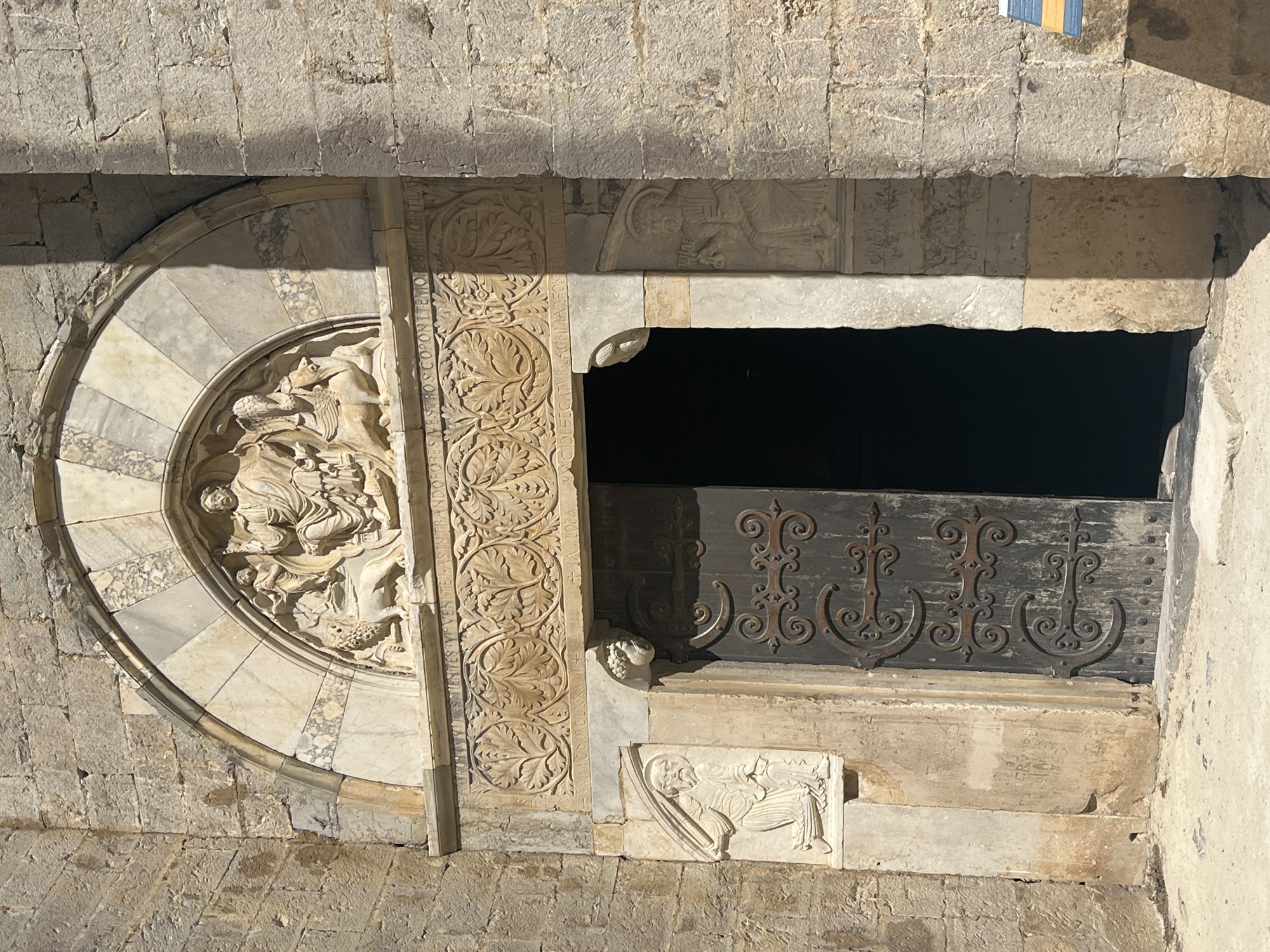 The image shows a close-up view of the carved stone portal of the Cathédrale Saint-Pierre de Maguelone, located in Villeneuve-lès-Maguelone, France. The portal features a semicircular arch with multiple carved layers. The uppermost layer displays a relief sculpture of a central seated figure, likely Christ, flanked by two other figures and two lions. Below this is a band of intricate floral carvings. Further down, on either side of the entrance, are carved figures. The left side depicts a figure holding a scythe, and the right side shows a figure with a halo, holding keys. The entrance itself is a large, dark wooden door with ornate wrought-iron hinges and decorative elements. The surrounding walls are constructed of rough-hewn stone blocks. The ground in front of the entrance is paved with concrete or stone. The lighting suggests it is a sunny day, with strong shadows cast by the architectural features.
