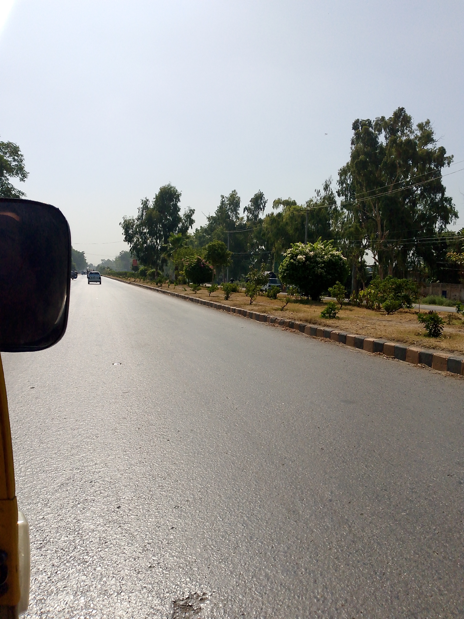 The image shows a view from a vehicle, likely a tricycle or autorickshaw, traveling on a paved road. The perspective is from the right side of the vehicle, with a partial view of a black side-view mirror in the foreground. The road is wide and appears to be a multi-lane highway or a major thoroughfare. There are cars visible on the road ahead, moving in the same direction.
The road is lined with trees on both sides, creating a green median and flanking the roadway. The trees are lush and varied in size and type, with some appearing to be eucalyptus. In the background, there are more trees and some signs of urban development, possibly buildings or walls, though they are indistinct due to the distance and haze. The sky is bright and overcast, suggesting it might be a hazy or partially cloudy day. The ground along the roadside appears dry and somewhat barren in places, with patches of grass and shrubbery. A distinct border made of alternating black and brown concrete blocks runs along the edge of the road. The overall impression is a scene of a typical road in a city with a lot of greenery.