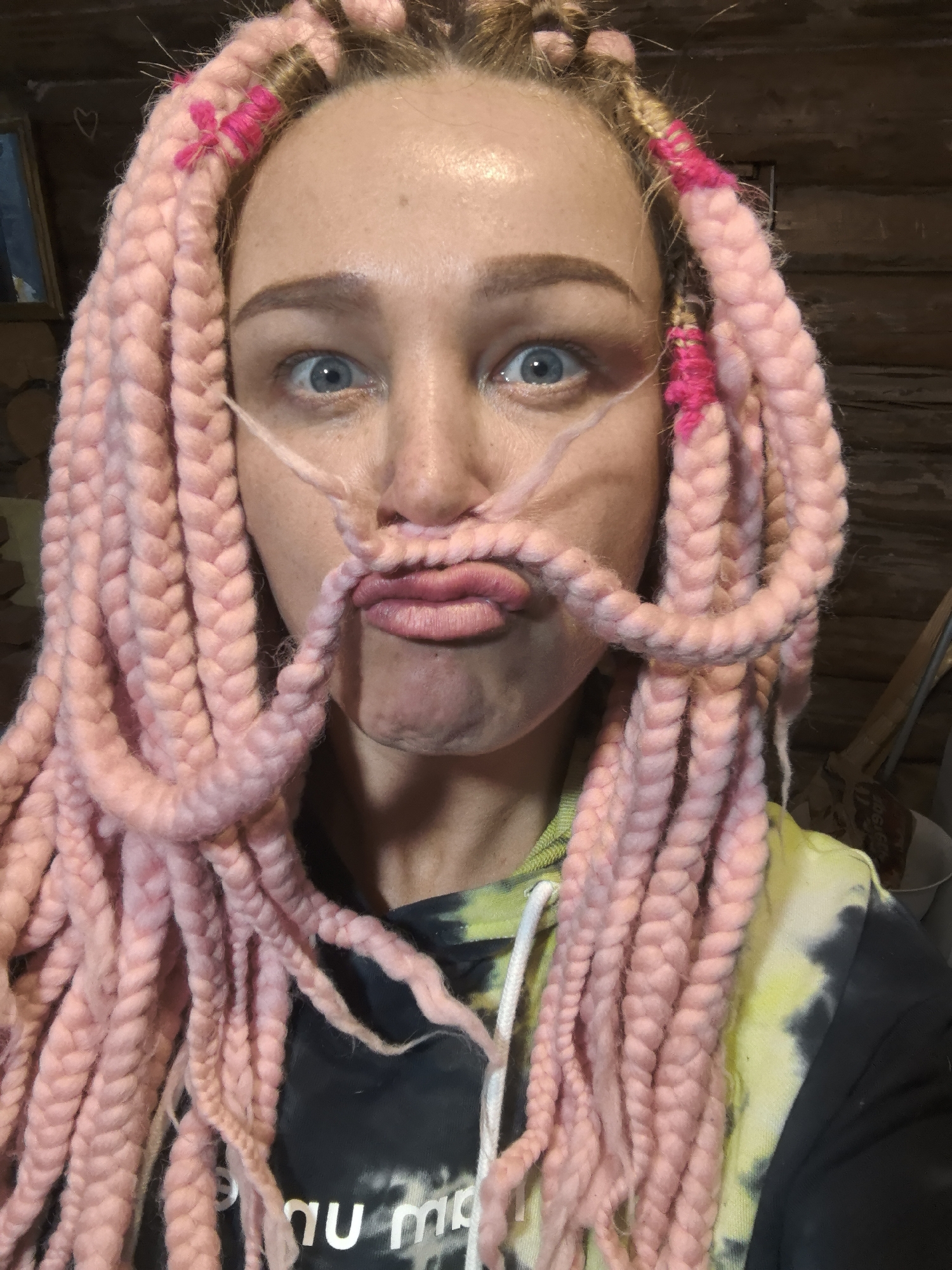 A woman with pink braids is posing for a close-up shot. She has pulled two of the braids up to her face to create a mustache effect. She is wearing a black hoodie with a yellow and green tie-dye pattern. The background is a blurred wooden wall.