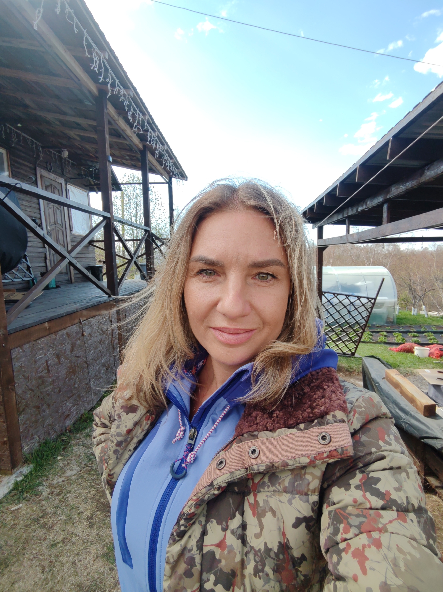 A woman with blonde hair stands outdoors wearing a camouflage jacket over a light blue zip-up hoodie. She is facing the camera with a slight smile. Behind her are two rustic wooden structures. One is a porch or small building with a peaked roof, and the other is a covered area open on at least one side. Beyond these structures is a greenhouse and a garden area with visible plants and some red bags scattered about. The sky is partly cloudy. The location is likely a rural or suburban area, possibly near Koryaki, Russia, based on the user's prompt.