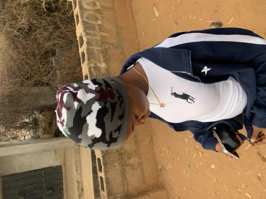 A person wearing a camouflage beanie, white t-shirt with a Polo emblem, and a navy blue track jacket is standing outdoors. They are holding a black object, possibly a phone, in their hand. The ground is tan or sandy. In the background, there is a low brick wall and some dry brush or shrubbery. The image is rotated so the person is sideways.