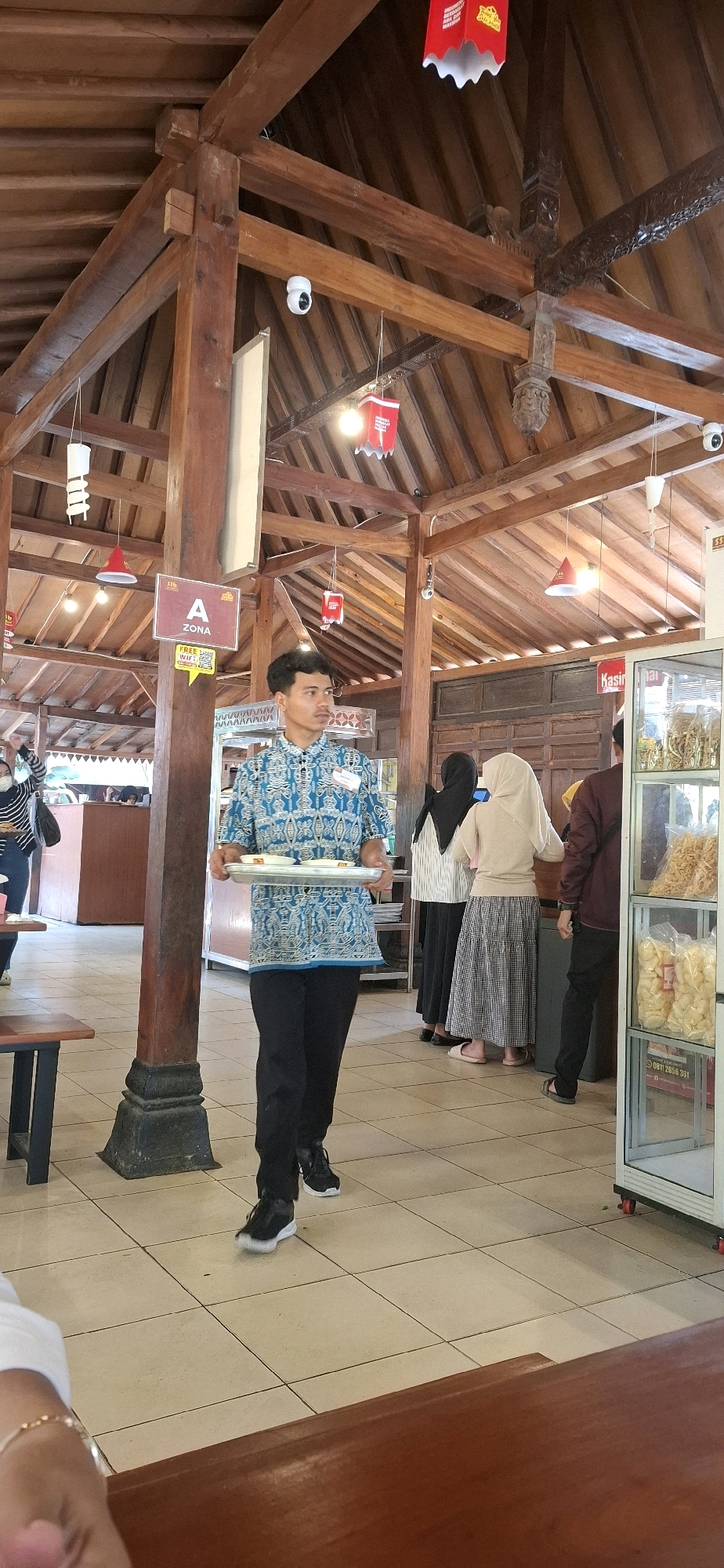 A young man in a blue and white patterned shirt and black pants walks through an indoor dining area, carrying a silver tray with several bowls of food. The architecture suggests a traditional Indonesian style, with exposed wooden beams and rafters. The walls are paneled wood, and the floor is tiled in a cream color. In the background, several other people are visible, including two women wearing headscarves and a man in a maroon shirt. A glass display case filled with snacks stands to the right. A sign above the man reads "A ZONA" with a "FREE WIFI" sticker below it. The scene appears to be a restaurant or food court in Colomadu, Indonesia, based on context provided.