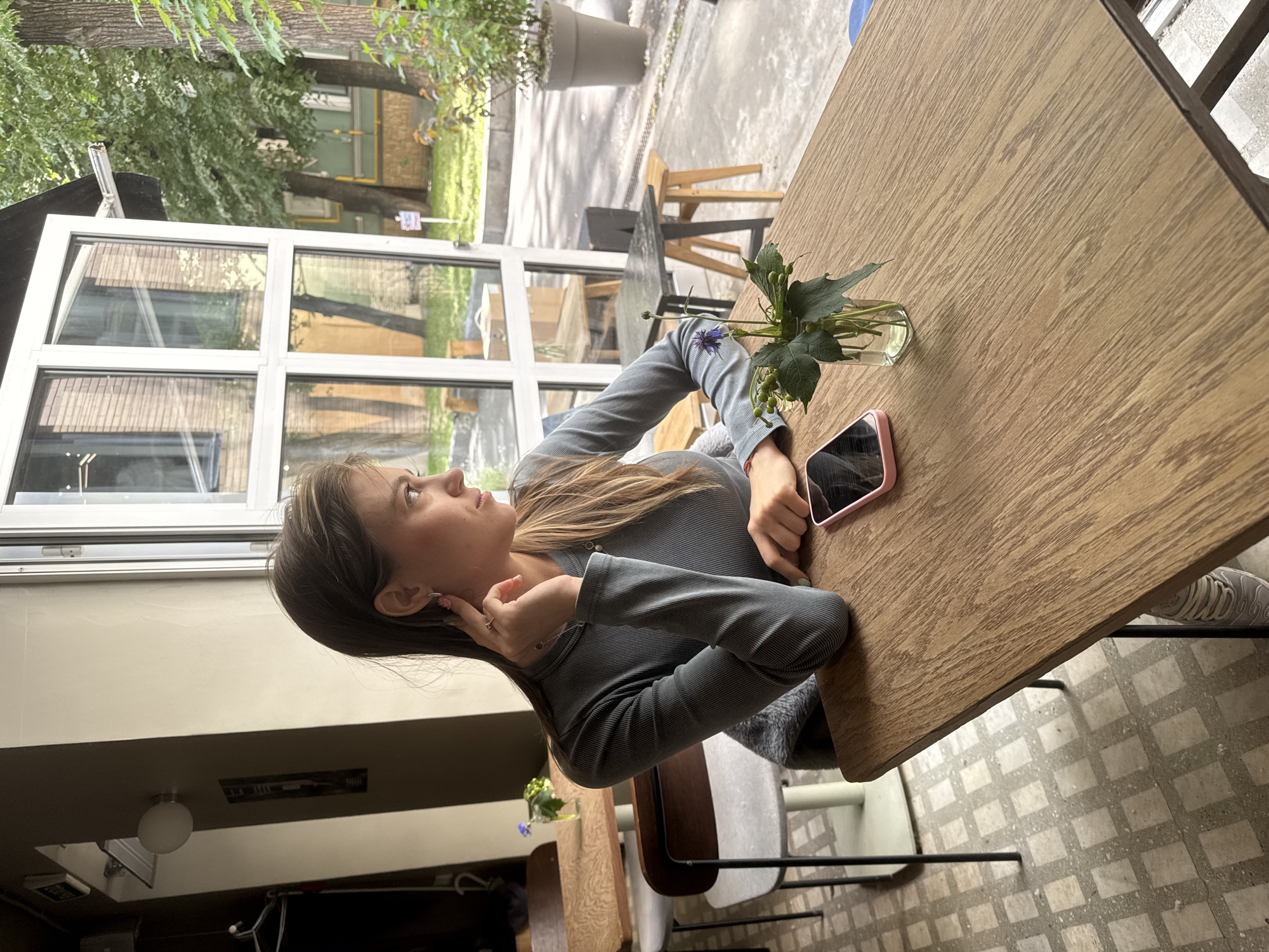 A young woman with long, blonde hair sits at a wooden table, looking to her left with a thoughtful expression. She is wearing a grey, long-sleeved, ribbed shirt. Her right hand is touching her ear, and a ring is visible on her left hand. A pink phone rests on the table in front of her, next to a small glass vase containing a few green stems and a single purple flower. The table is set in what appears to be an outdoor cafe or a room with large windows overlooking a green, leafy area with trees and buildings. The floor is tiled with a grey and white checkered pattern. The lighting suggests it is daytime, possibly overcast. The scene conveys a sense of quiet contemplation or a moment of pause.