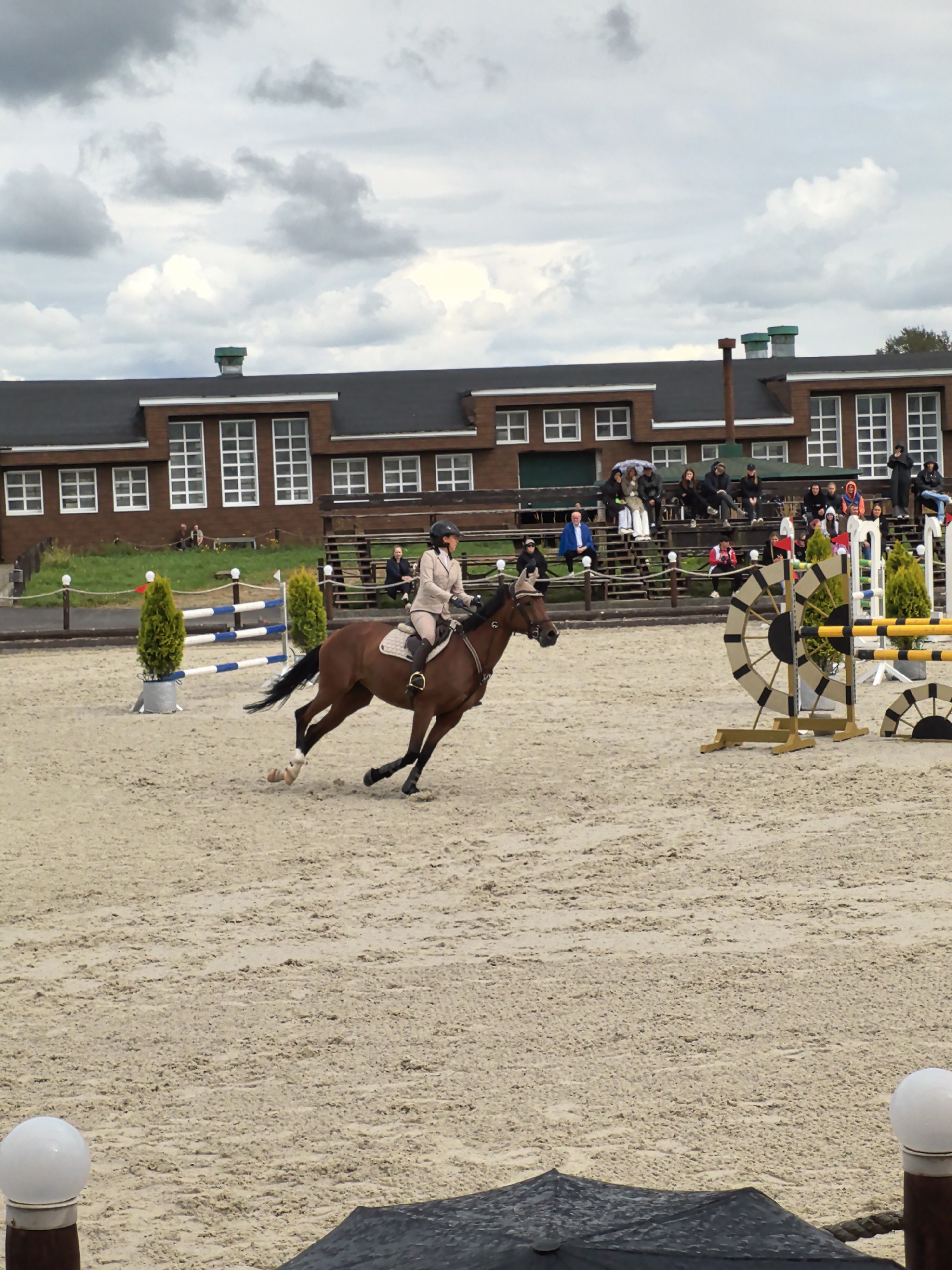 An equestrian competition is underway at an outdoor arena. A rider on a brown horse is in mid-gallop, navigating the sandy track. The rider is wearing a tan riding jacket and helmet. In the background, a long, multi-story building with a dark roof and numerous windows stands against a cloudy sky. Spectators are seated on bleachers in front of the building. Various jumps and obstacles are set up within the arena, including a large, decorative circular jump. Two potted evergreen trees are placed near a jump pole. The ground of the arena is covered in fine sand. Two white spherical lights are visible in the foreground. The location is identified as a farm in Russia.