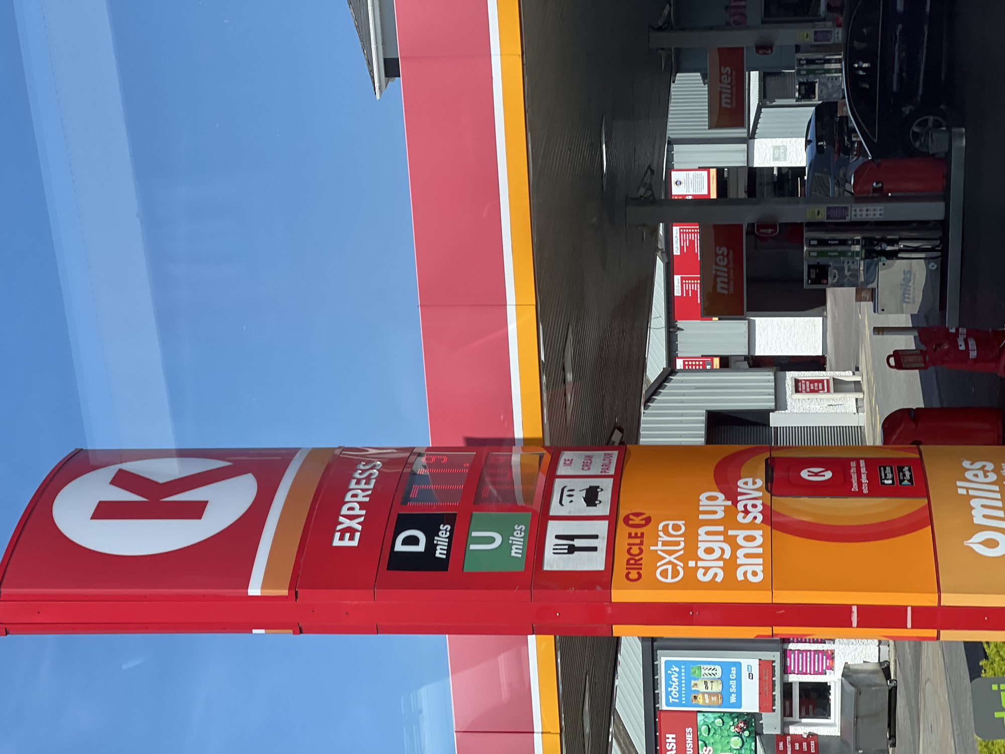 The image shows a Circle K Express gas station in Letterkenny, Ireland, on a bright, sunny day. A prominent red and white Circle K sign stands tall, displaying the brand's logo and "EXPRESS" text. Below the main sign, a digital display shows fuel prices. Further down, icons represent dining, a car wash, and an ice cream parlor. A large orange sign advertises "Circle K Extra sign up and save" with a QR code for downloading the app. The gas station canopy, painted in red and yellow, covers multiple fuel pumps. Cars are visible, with one black car parked near the pumps. In the background, buildings and a grey trash bin are present. A sign for "Tobin's Letterkenny" is also visible on the left side of the image.