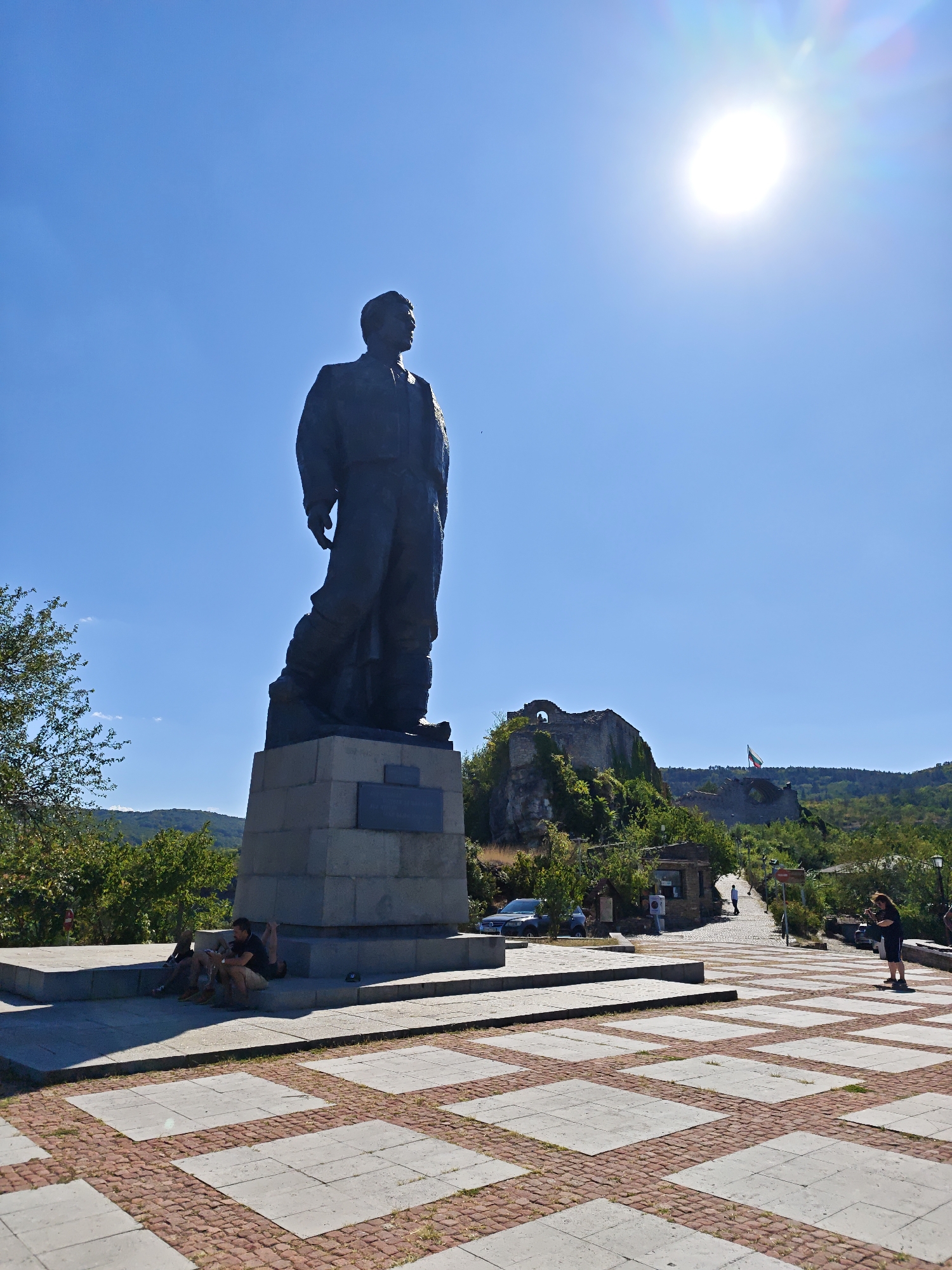 A monumental bronze statue of a man in uniform stands on a stone pedestal in a public square. The statue is viewed from a low angle, with the bright sun in the upper right corner of the frame, causing lens flare. In the background, a castle ruin is visible on a hilltop, with a Bulgarian flag flying above it. A few people are seated on the steps of the pedestal, while others are walking in the square. The square is paved with large square tiles and red brick. Lush green trees and rolling hills are visible in the distance. The overall scene suggests a tourist destination on a clear, sunny day. This is likely the monument to General Voivode Vasil Levski in Lovech, Bulgaria, given its prominent location and the historical context of the castle ruins.