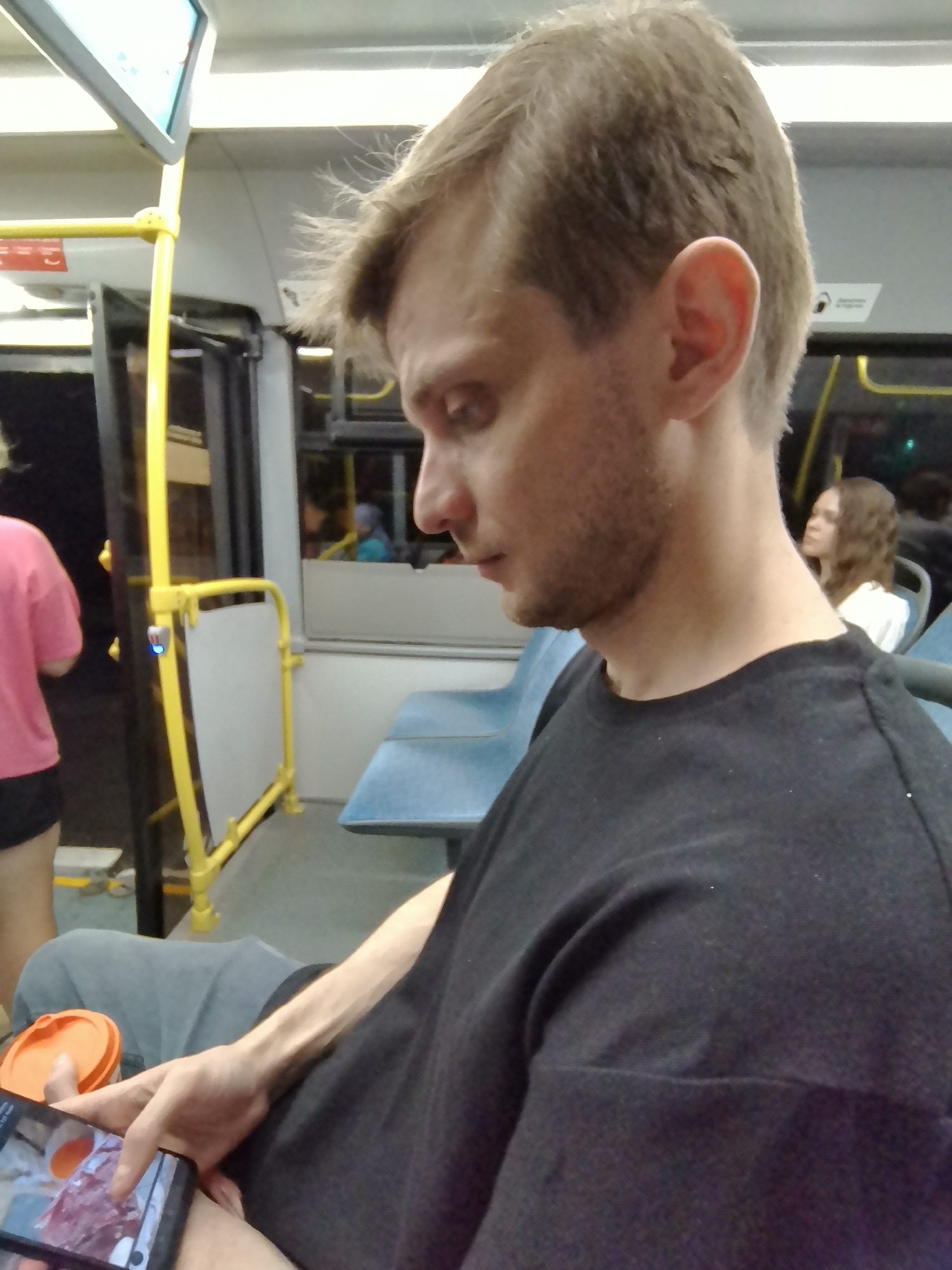 The image shows a young man with short brown hair and a beard, wearing a black t-shirt, sitting on a bus. He is looking down at his phone, which he is holding in his left hand. An orange cup with a lid is placed next to his phone. In the background, there are blue bus seats and a yellow handrail. Further back, a young woman with brown curly hair is seated. The scene appears to be inside a public bus in Krasnogorsk, Russia.