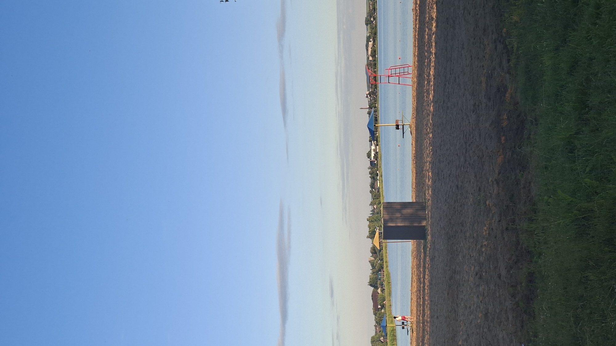 The image depicts a serene beach scene by a body of water, likely a lake or river, with a town or village visible on the opposite shore. The foreground is dominated by a sandy beach with sparse green grass at the very bottom edge of the frame. In the middle ground, there are several beach amenities: a grey changing booth or structure, a blue umbrella with a bench beneath it, and a red lifeguard chair with a ladder. A person wearing red shorts and a white top is standing on the sand near the changing booth, facing away from the camera. The water is calm and reflects the clear blue sky, which has a few wispy clouds. The distant shoreline features a line of trees and buildings, suggesting a populated area. The lighting indicates it is either early morning or late afternoon, with long shadows cast on the sand. The overall atmosphere is peaceful and recreational. This scene is consistent with a public beach area in or near the city of Smolevichi, Belarus, known for its numerous lakes and recreational spots.