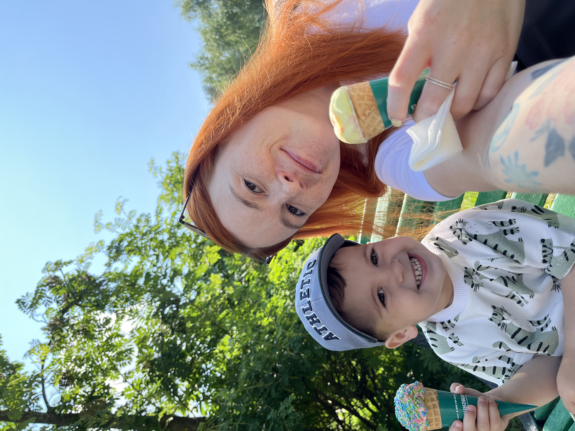 This is a close-up, selfie-style shot of a woman and a young boy with bright red hair and pale skin, sitting outdoors on a sunny day, possibly in a park in the city of Chaikovsky, Russia, as indicated by the information provided. The woman, positioned on the right, is looking towards the camera with a smile, holding a small ice cream cone with yellow ice cream and a green wrapper. She has freckles and her long, red hair is visible, with sunglasses perched on top of her head. She is wearing a white top. The boy, on the left, is wearing a grey baseball cap with "ATHLETIC" written on it and a white t-shirt with a pattern of grey cats. He is smiling widely, showing his teeth, and holding a different ice cream cone topped with pink ice cream and colorful sprinkles. The background consists of lush green trees and a clear blue sky, suggesting a warm, pleasant day. The overall atmosphere is casual and happy, capturing a moment of enjoyment between the two individuals.