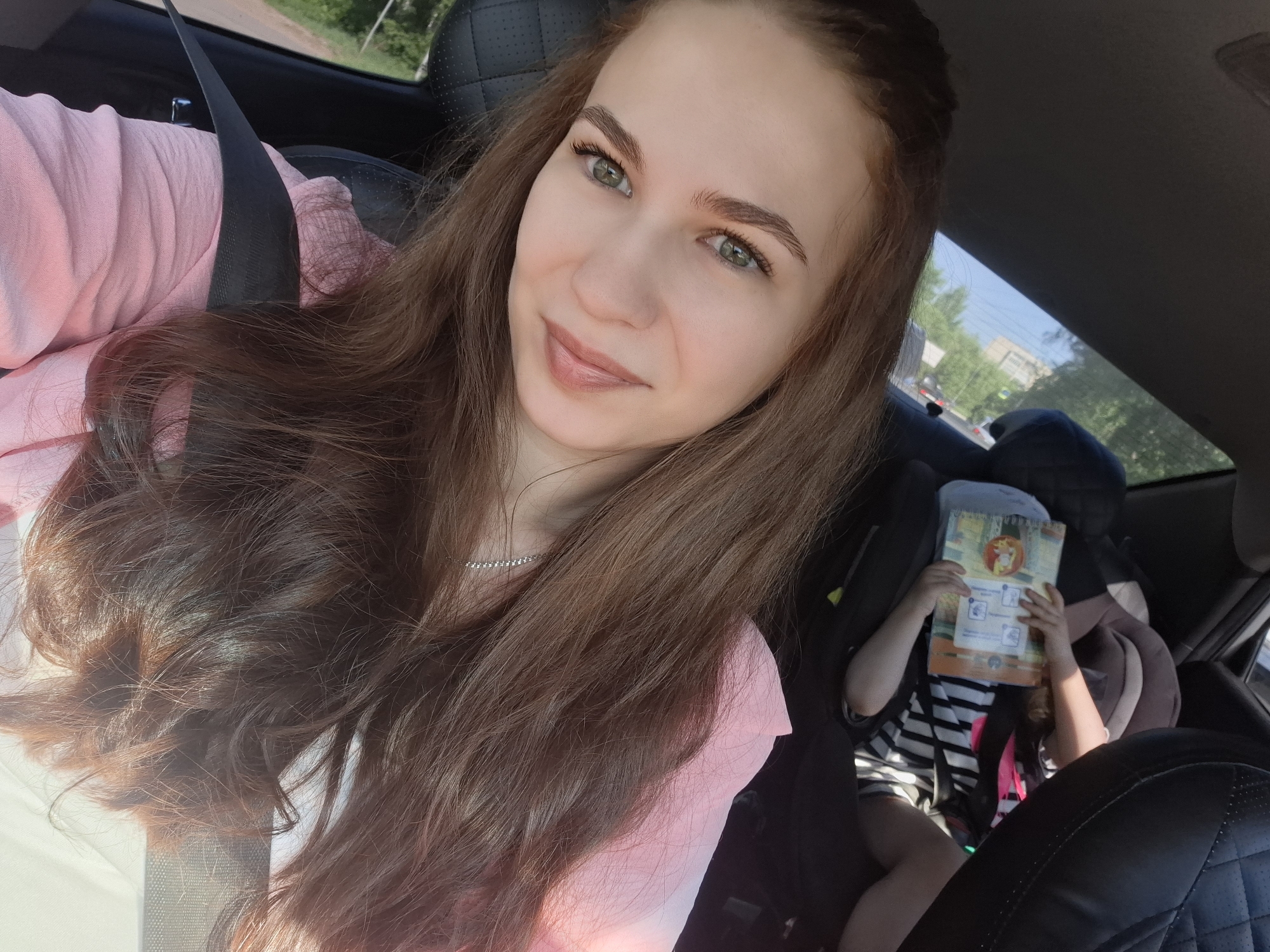 A selfie shows a woman in the front passenger seat of a car, wearing a pink top and a seatbelt. She has long brown hair, green eyes, and light skin. In the back seat is a child in a car seat, holding a book in front of their face. The child is wearing a black and white striped shirt and is buckled into the car seat. Outside the car window, there is a view of buildings and trees, suggesting a location within a city. Based on the metadata, the location is Chaikovsky, Russia. The woman appears to be taking a selfie with the child in the back.