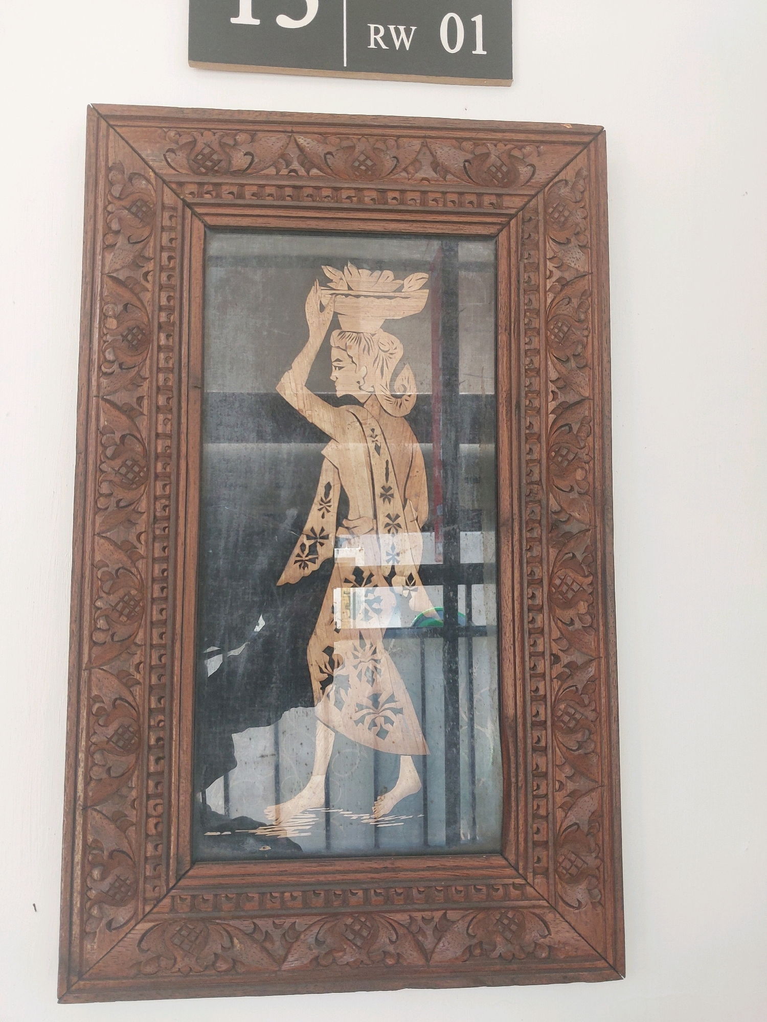 This image shows a framed artwork hanging on a white wall. The artwork features a silhouette of a woman carrying a basket of objects on her head. The woman is depicted in profile, walking from left to right, and appears to be wearing a dress with floral patterns. The frame is made of intricately carved wood. Above the artwork, a sign with "15 RW 01" is visible. The sign is dark gray with white lettering. The setting is likely indoors, given the white wall and the presence of a framed artwork. The overall aesthetic suggests a decorative piece, possibly in a residential or public space in Malang, Indonesia, as indicated by the number "15" potentially referring to a house number or room in that city.
