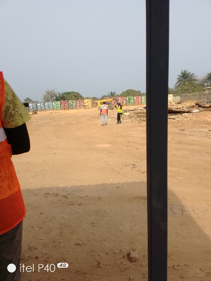 This image depicts an outdoor industrial or storage yard in Hoche, Nigeria. The sky is clear and light-colored. The ground consists of unpaved, dry, light brown soil with scattered small rocks.
In the immediate foreground on the left, a person is partially visible, wearing an orange high-visibility vest over a light green patterned shirt and dark sleeves. A dark vertical metal pole or beam partially obstructs the right side of the foreground.
In the midground, two individuals are present. One person, positioned to the left, is seen from behind, wearing a red-orange high-visibility vest over a light shirt and light-colored pants. To their right, another person wears a bright yellow high-visibility vest over dark clothing and has their right arm raised in a gesturing motion, directed towards the first individual.
In the background, multiple rows of stacked shipping containers of various colors (blue, green, red, brown) are arranged horizontally across the scene. A large yellow piece of heavy machinery, possibly a telehandler or forklift, is positioned to the left of the two midground individuals and in front of the containers. To the far right, a pile of debris, including what appears to be wood and metal, is visible. Beyond the containers and debris, several green trees, including palm trees, are present under the distant horizon.