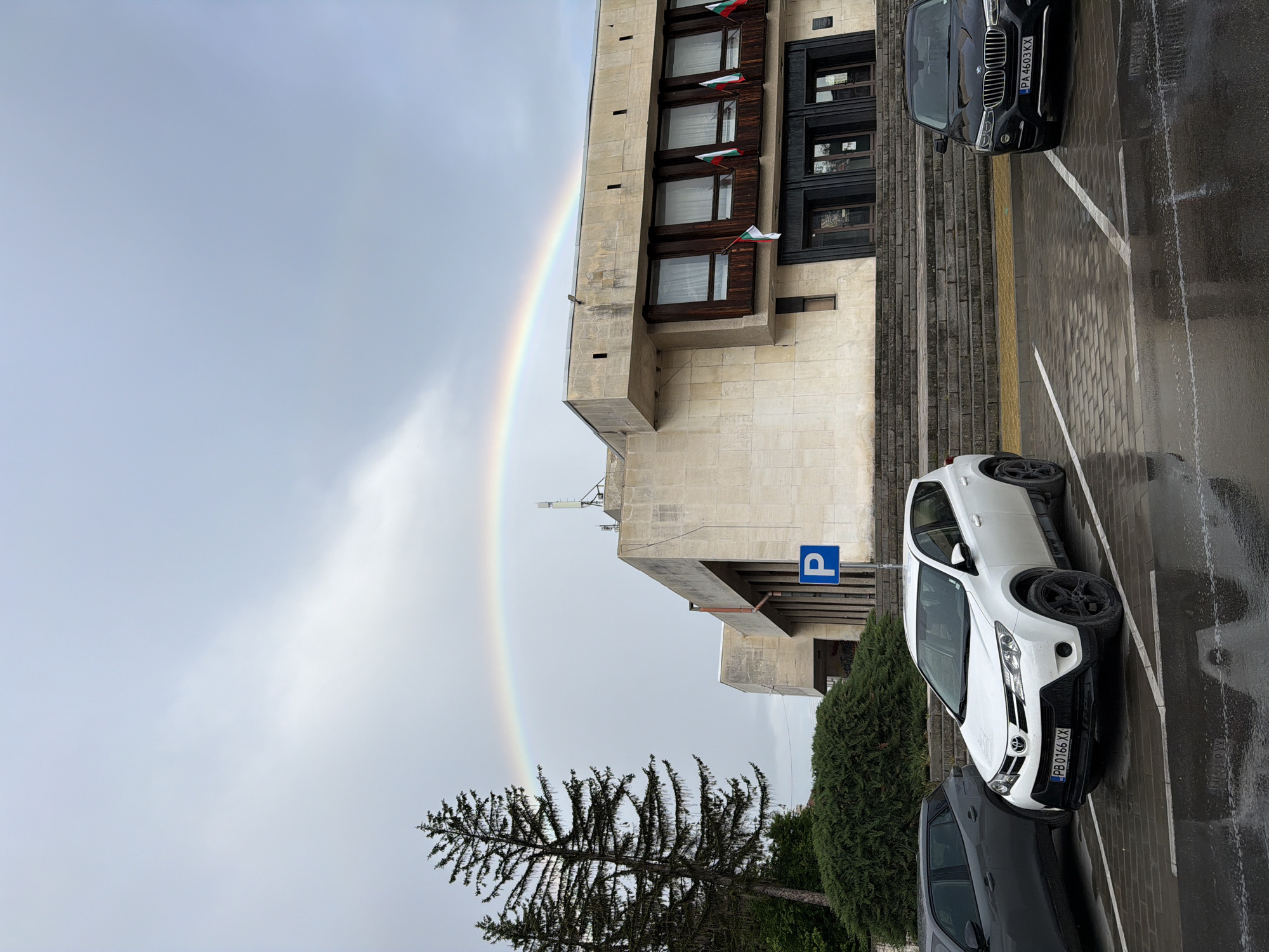 The image depicts a rainbow ending near a building in Panagyurishte, Bulgaria. The building appears to be a government or municipal building, as it displays multiple Bulgarian flags from its windows. The architecture of the building is modern, with a facade of beige or light-colored stone or concrete. A "P" sign (parking sign) is visible near the building.
In the foreground, there are parked cars on a wet surface, suggesting it has recently rained. One of the cars is a white SUV with a license plate "PB 0166 XX". A dark-colored car is parked beside it. A tree is situated near the edge of the frame. The sky is overcast, and the rainbow arches across the sky, terminating near the left side of the building.